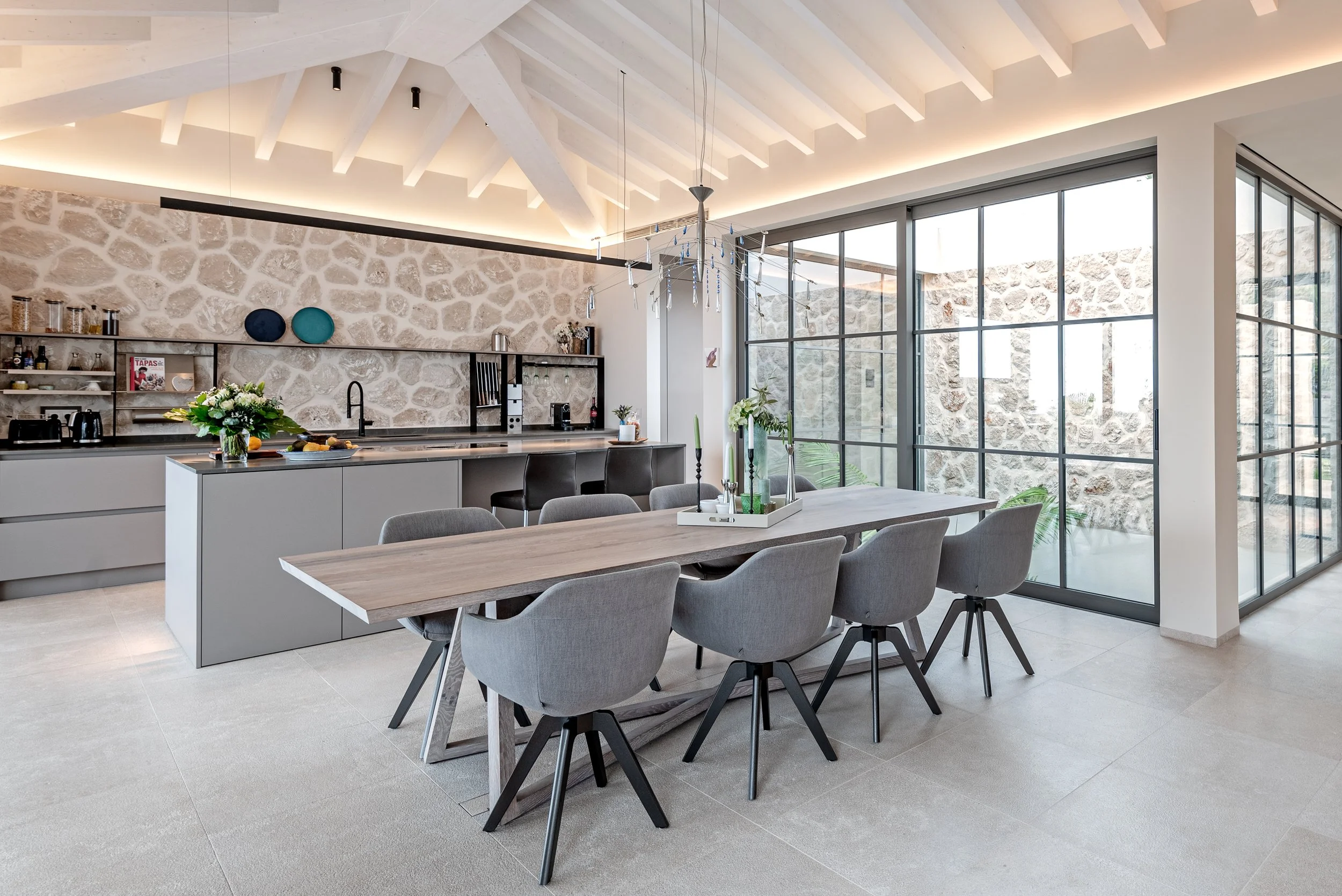 Interiores de una cocina moderna con una mesa de comedor, sillas grises y una pared de piedra decorativa por encima de la encimera, grandes ventanas de vidrio que dejan entrar luz natural y plantas decorativas.