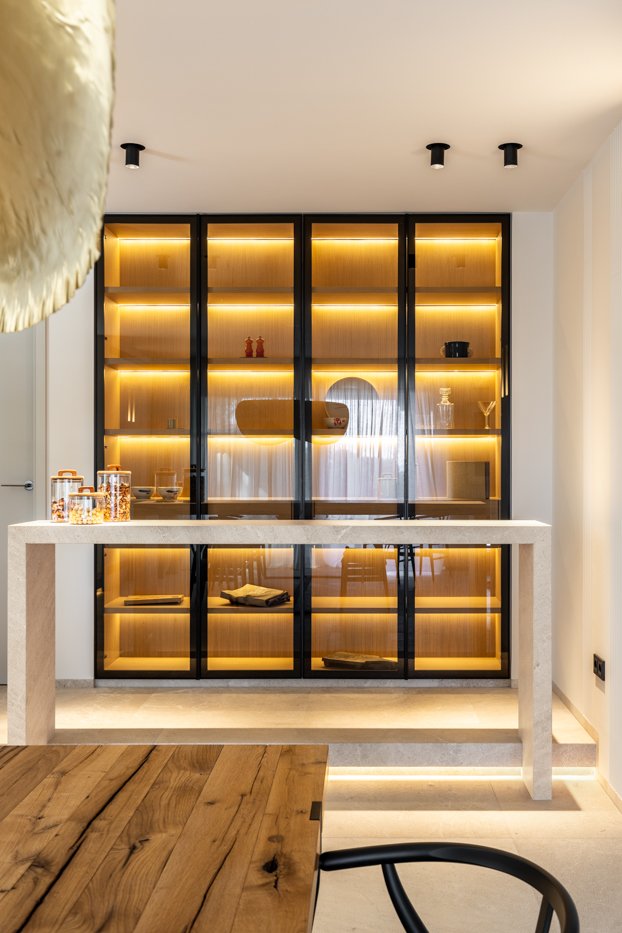 Vitrina moderna con puertas de vidrio negro y estantes con iluminación cálida en una pared blanca, decorada con objetos en exhibición.