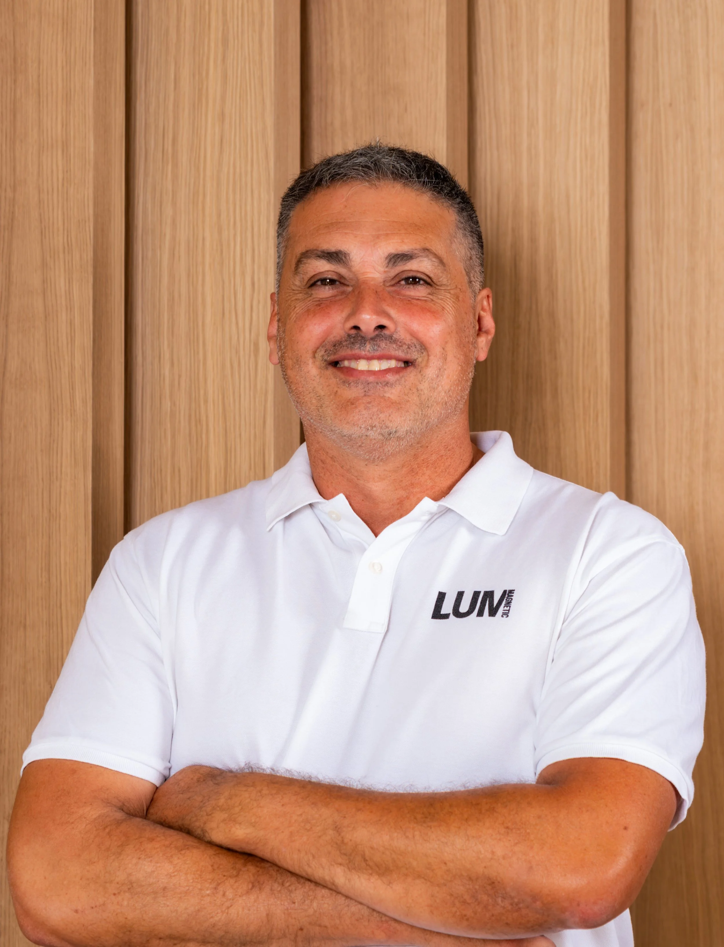 Hombre sonriendo con brazos cruzados, lleva una camiseta blanca con logotipo LUM, fondo de paneles de madera.