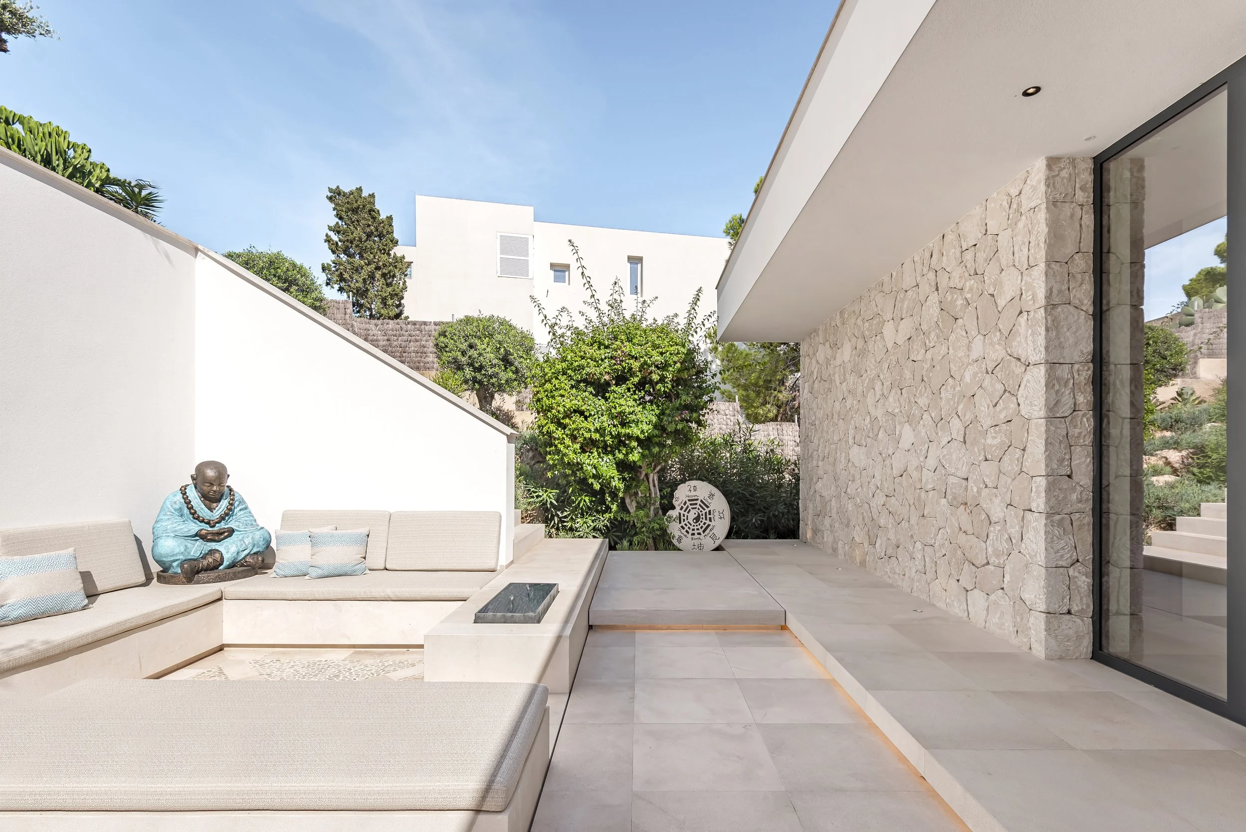 Espacio exterior moderno con mobiliario de color beige, una estatua de un monje en color azul, plantas verdes y un muro de piedra en uno de los laterales, bajo un cielo despejado.