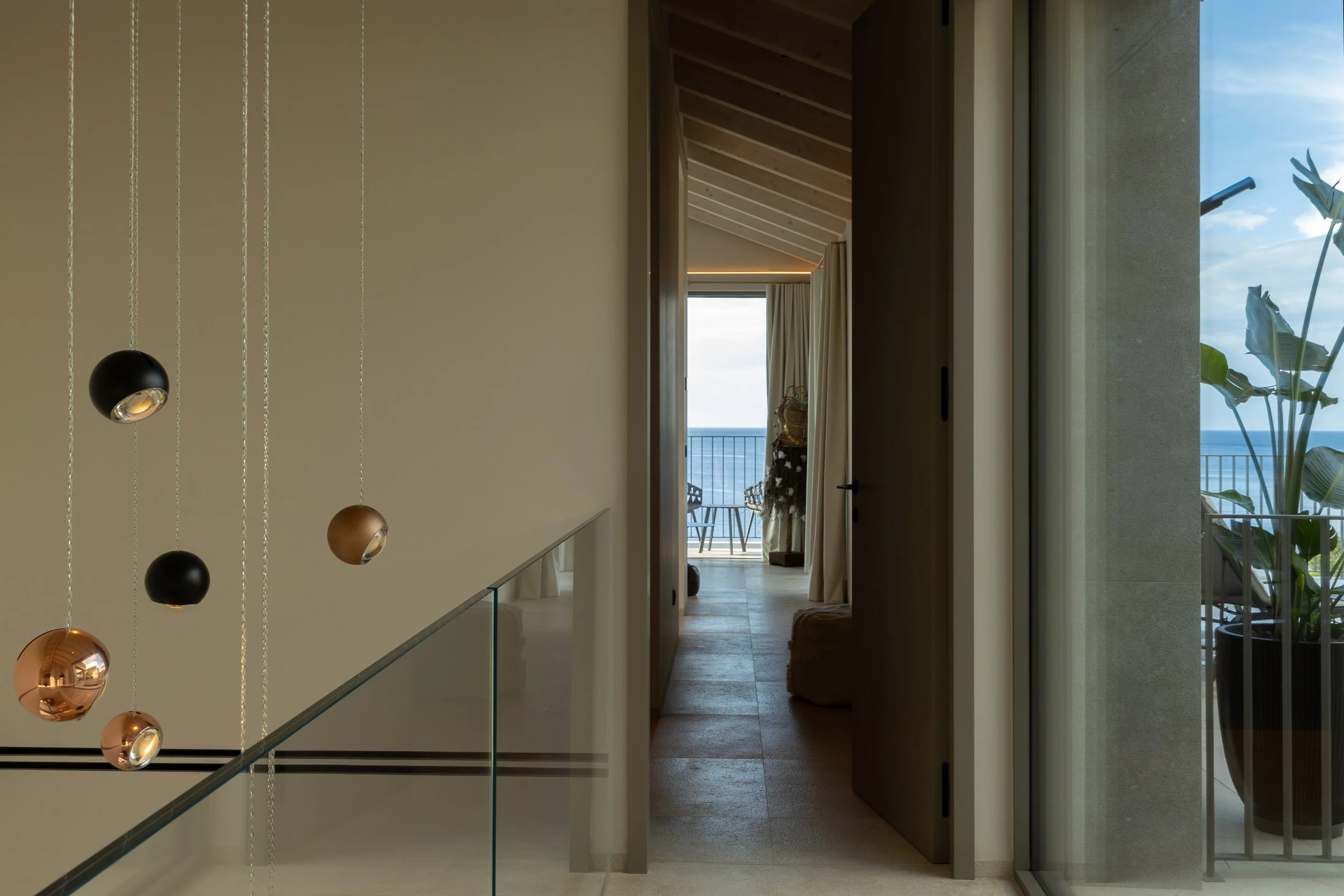 Interior de una casa moderna con vista al mar, balcón y plantas, con puertas abiertas y muebles en fondos.
