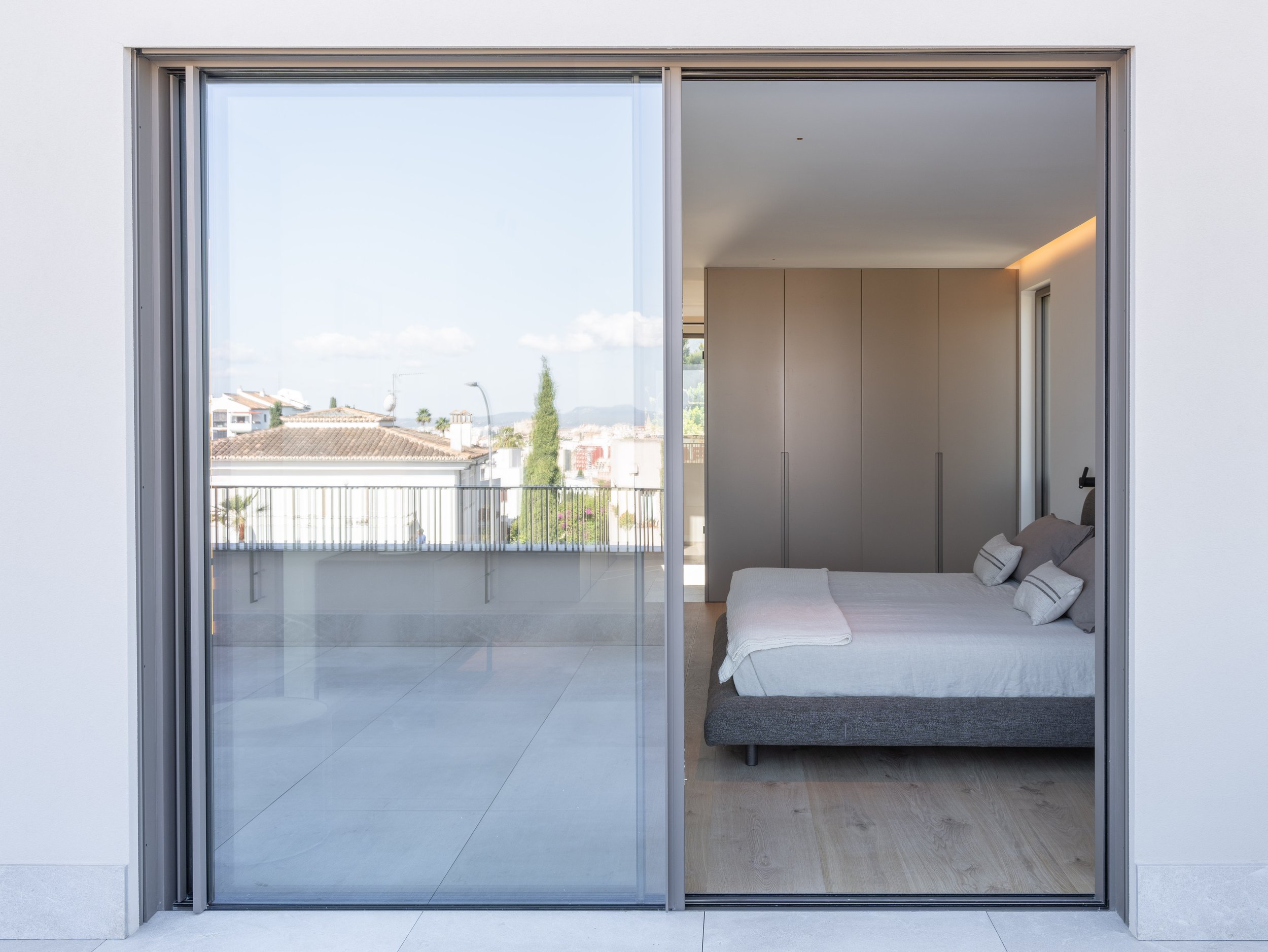 Vista de un dormitorio moderno con una cama gris, almohadas blancas con franjas negras, un armario beige, y una gran ventana de piso a techo con vista a un vecindario y cielo despejado.