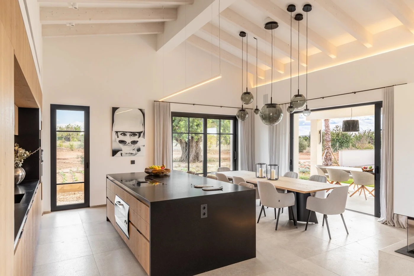 Cocina moderna y luminosa con isla de madera y sillas blancas, comedor con mesa de madera y sillas blancas, grandes ventanas y puertas corredizas con vista al exterior, decoraciones minimalistas y cuadros en las paredes.