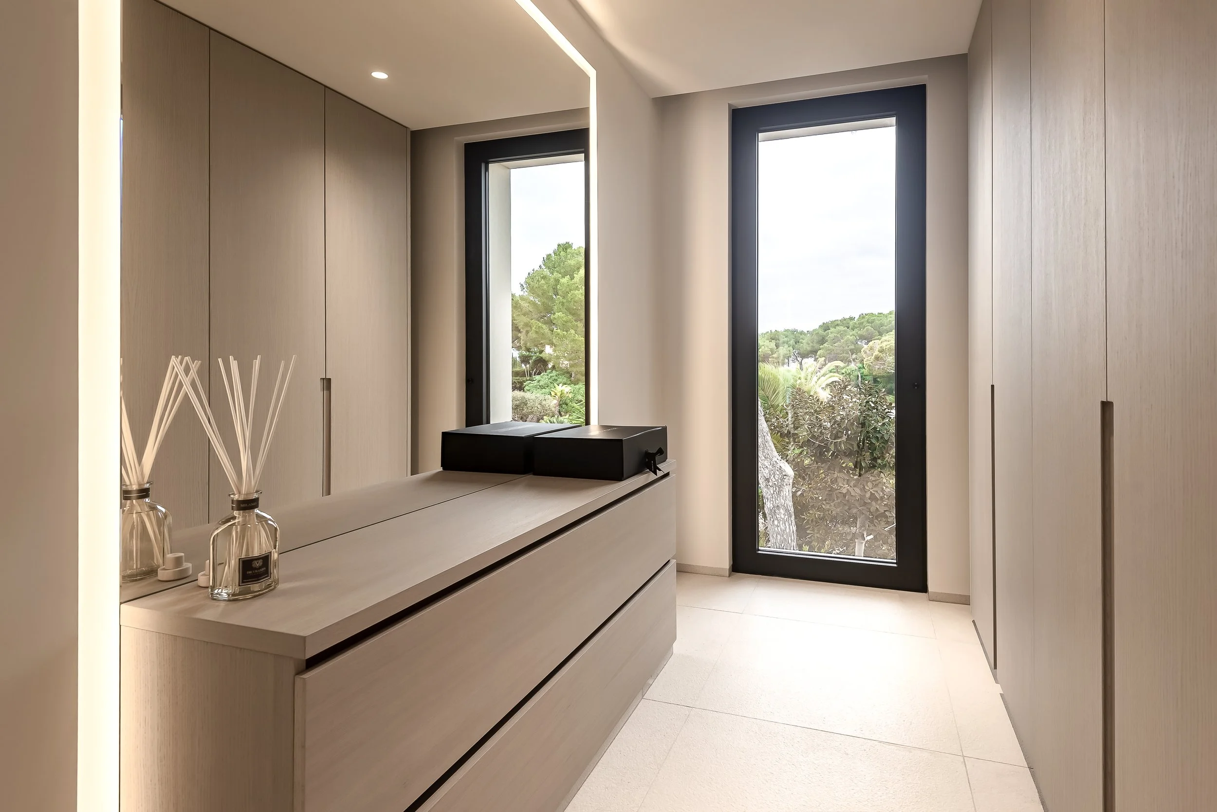 Interior de una habitación con ventanas grandes que permiten la vista del entorno natural exterior, con mobiliario minimalista de color claro y decoración sencilla.