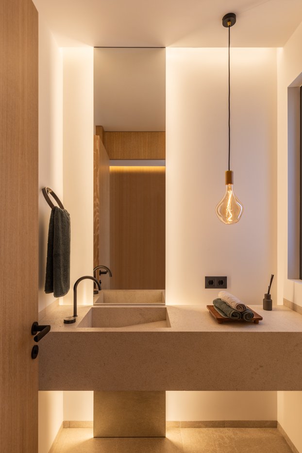Lavabo de baño moderno con encimera de piedra, un grifo negro, una toalla gris colgada en un gancho, un espejo grande, un colgante con luz de estilo vintage, y objetos decorativos de color neutro.