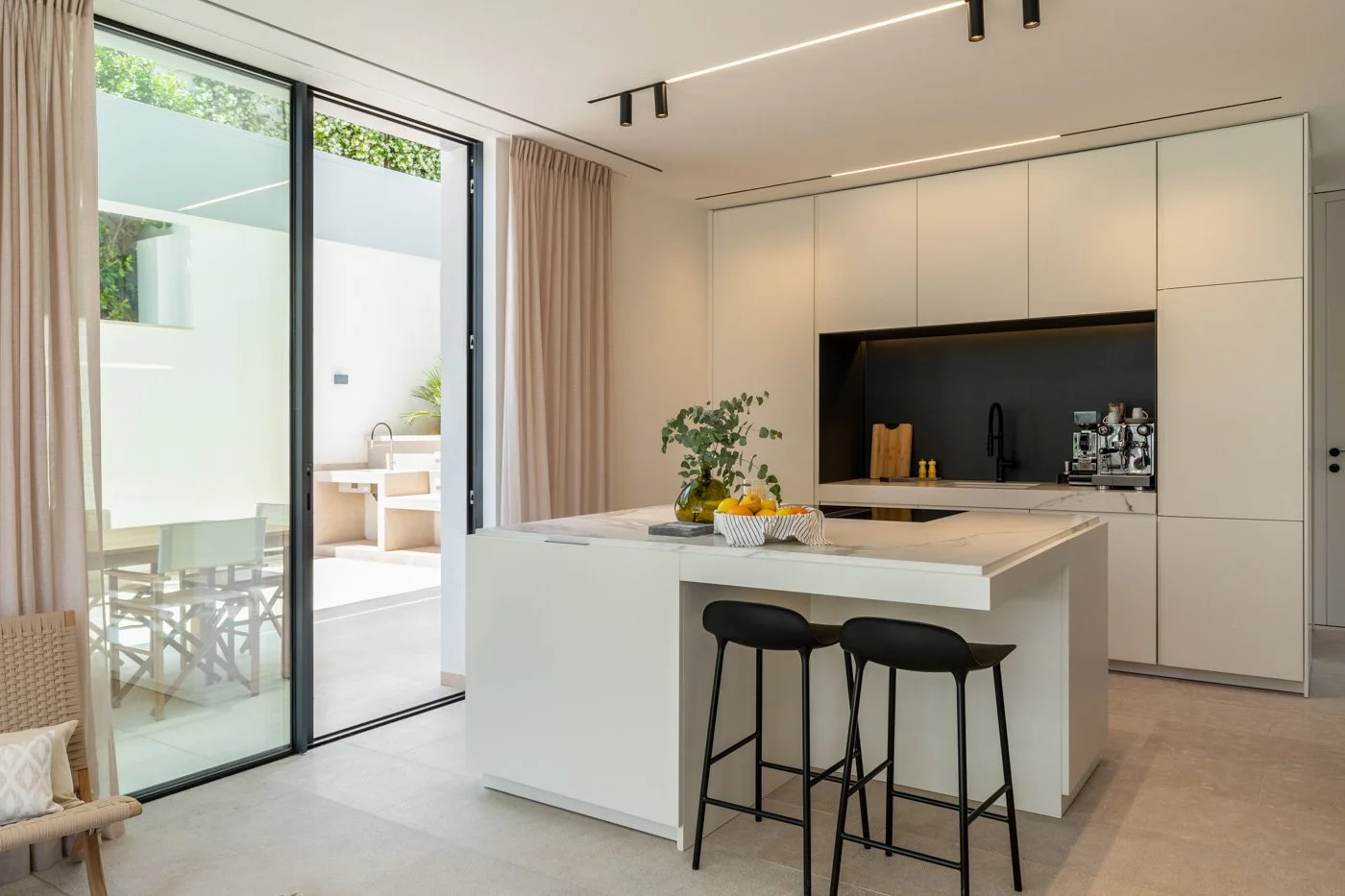 Cocina moderna y minimalista con isla blanca, dos taburetes negros, armarios blancos y una puerta de vidrio que conduce a un patio exterior con mobiliario.