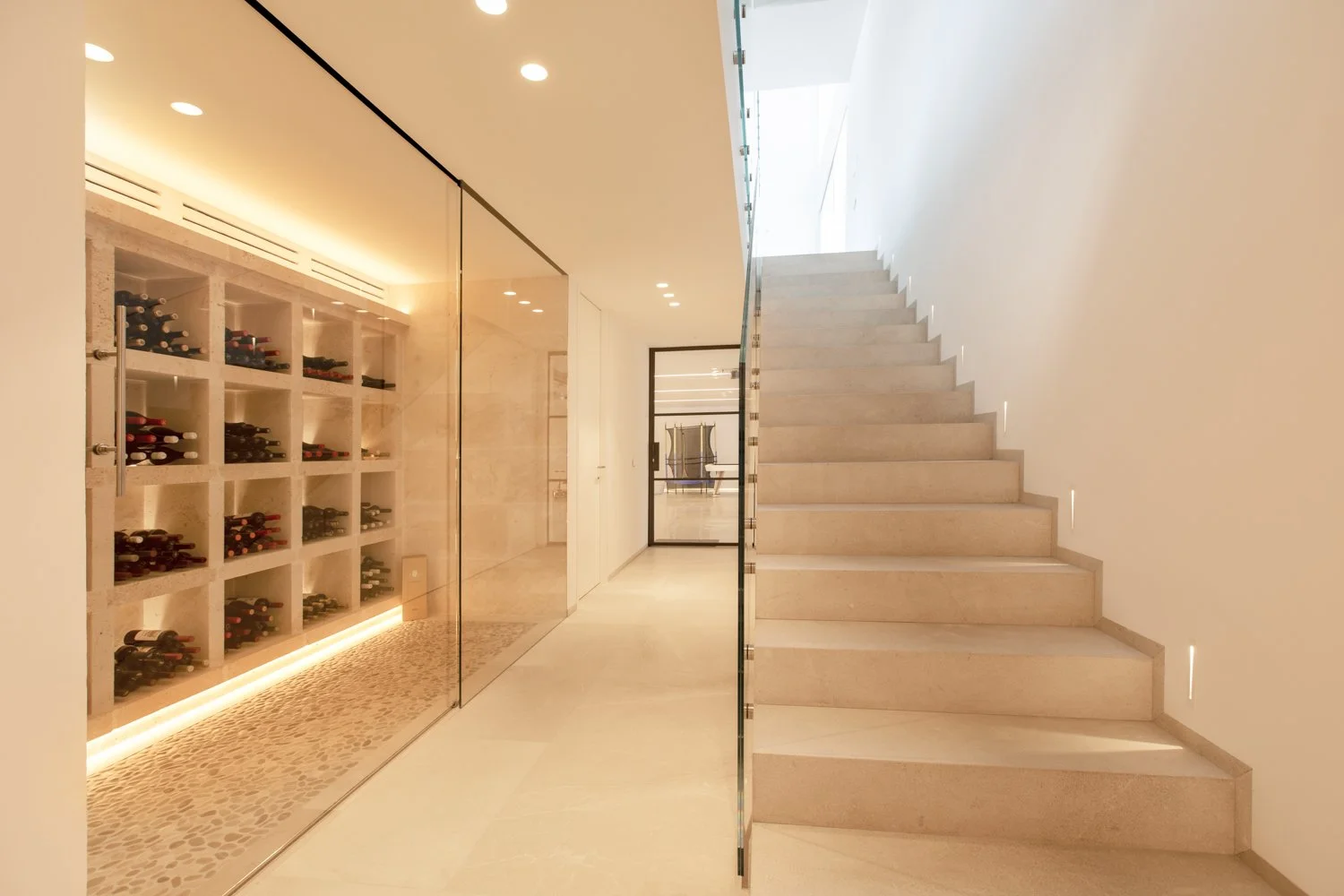 Pasillo con pared de cristal a la izquierda que muestra una bodega de vinos y una escalera blanca a la derecha.