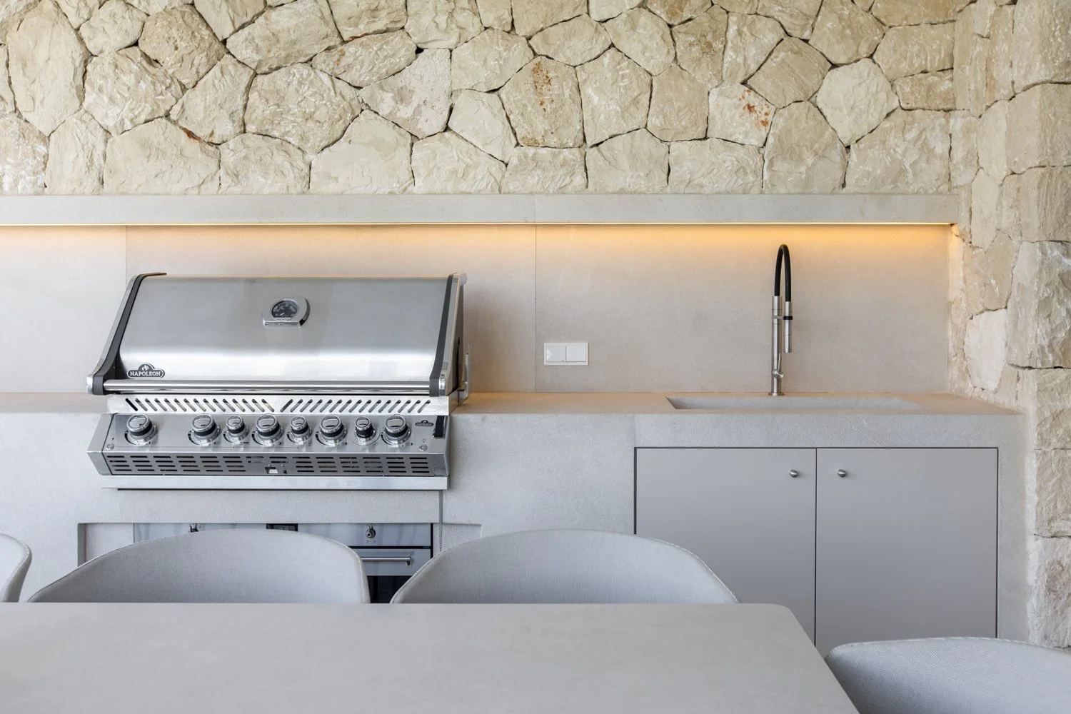 Cocina moderna con fachada de piedra, estufa de gas metálica, fregadero con grifo negro y armario inferior, mesas y sillas blancas.