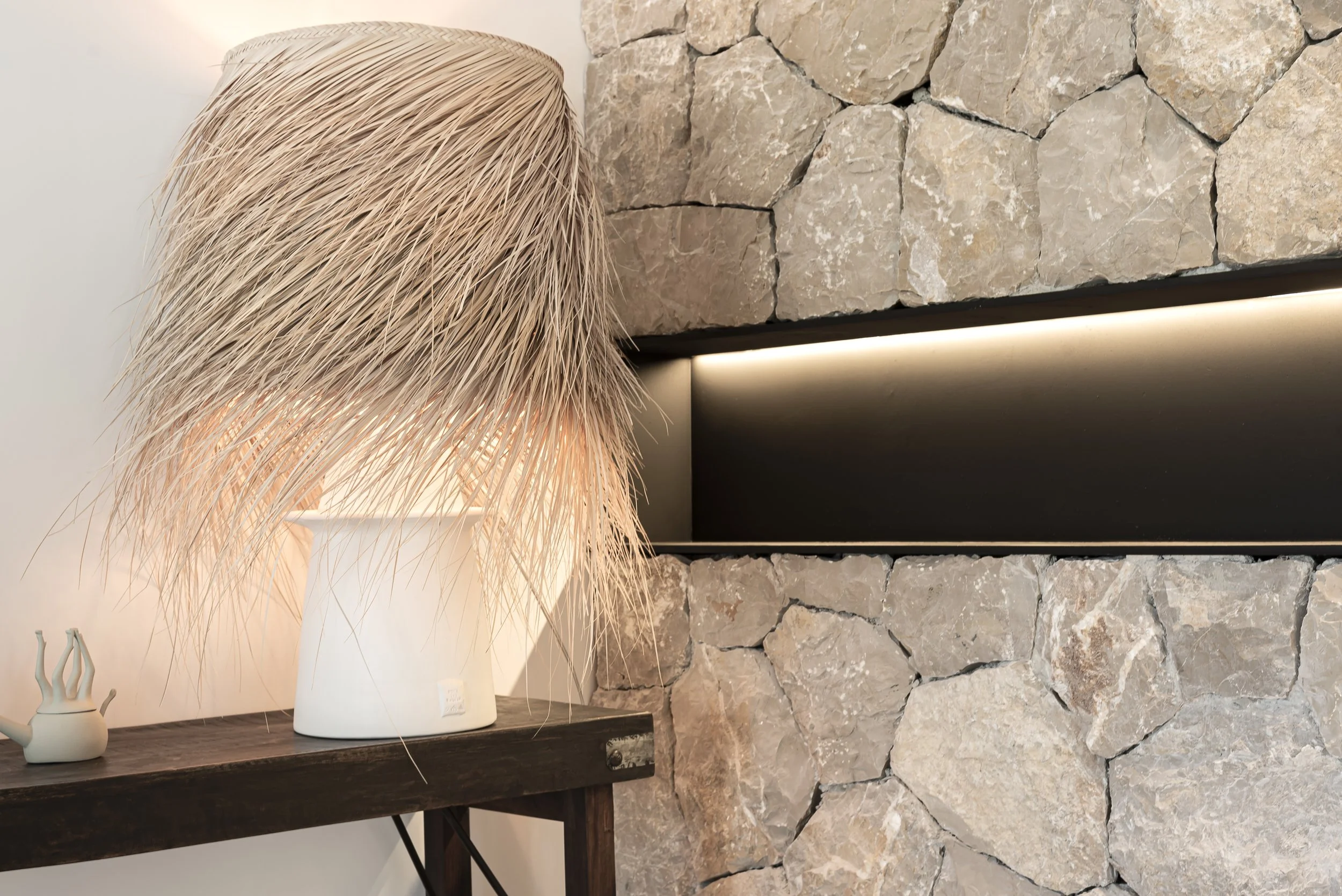 Lámpara de madera decorativa sobre una mesa de madera junto a una cerámica pequeña en una sala con pared de piedra