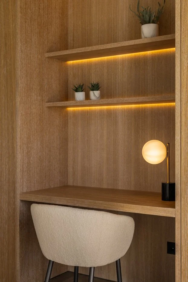 Estudio o rincón de trabajo con estantes de madera, pequeñas plantas en macetas blancas, una lámpara de luz cálida y una silla beige con patas negras.