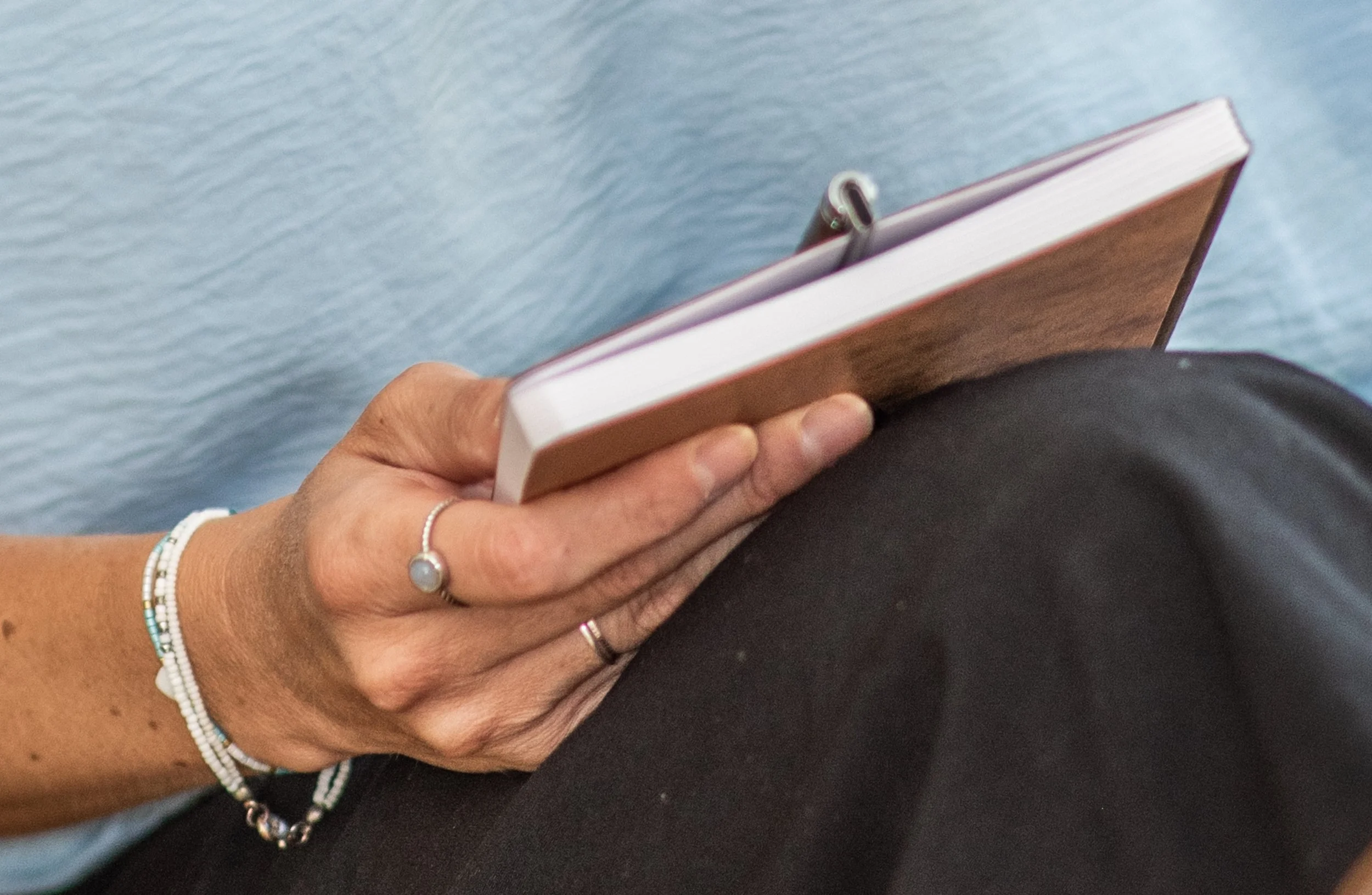 Nahaufnahme einer Hand, die ein Buch liest, mit mehreren Armbändern und Ringen, vor einem blauen Hintergrund.