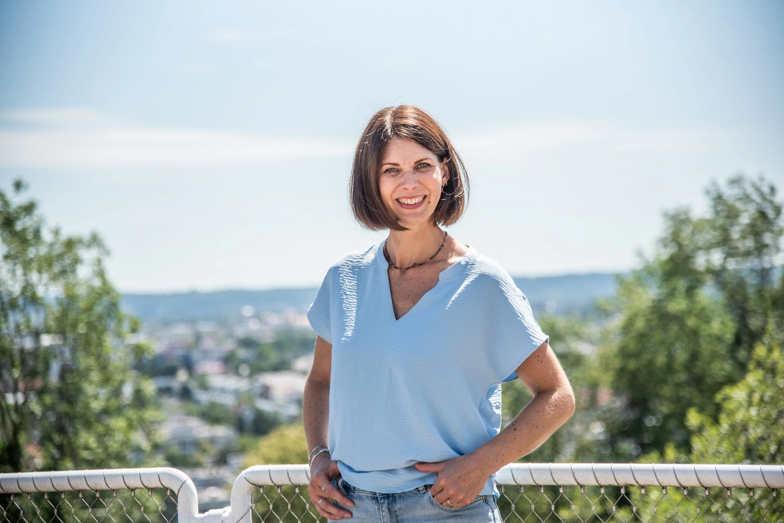 Eine lächelnde Frau mit kurzer brauner Haar, trägt ein hellblaues T-Shirt und eine Halskette, steht vor einer Metallabsperrung im Freien bei sonnigem Wetter, mit einer Stadt im Hintergrund und Bäumen auf beiden Seiten.