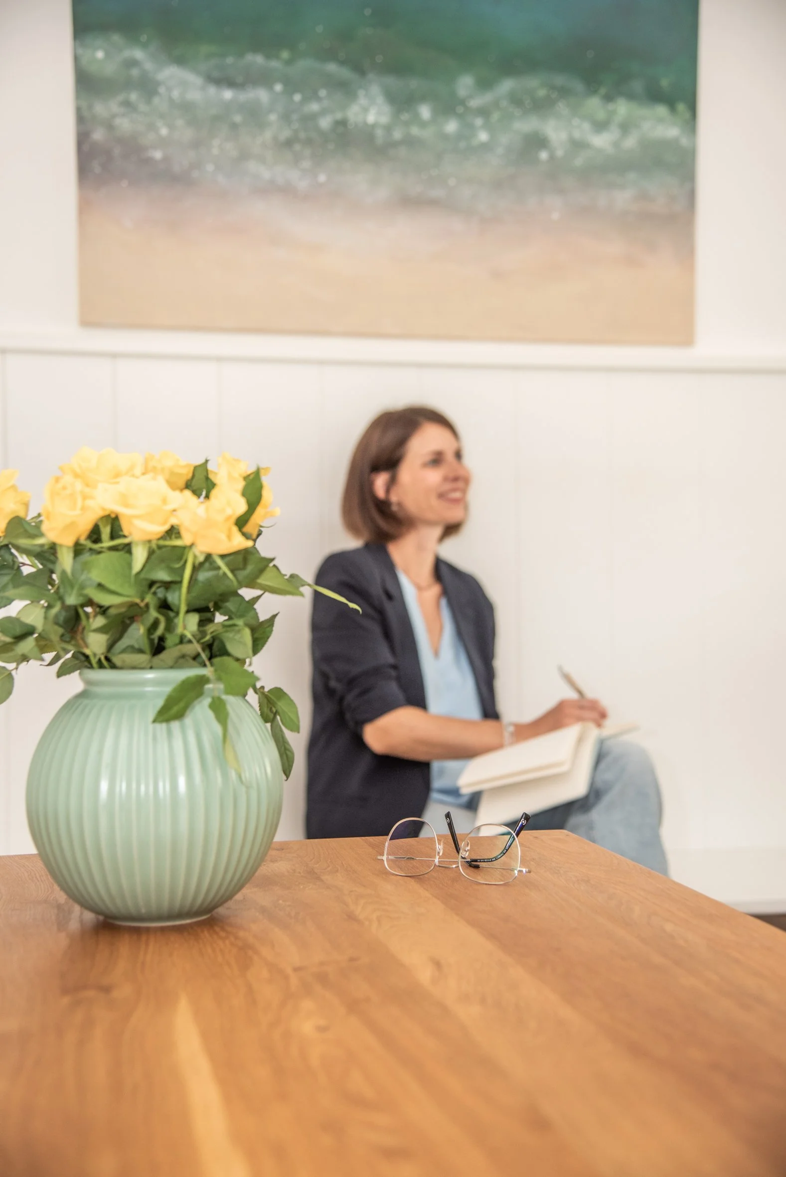 Blick auf eine Vase mit gelben Blumen auf einem Holzschreibtisch, im Hintergrund eine Frau mit braunen Haaren, die sitzt, liest und notiert, in einem Raum mit weißer Wand und Kunstwerk an der Wand.