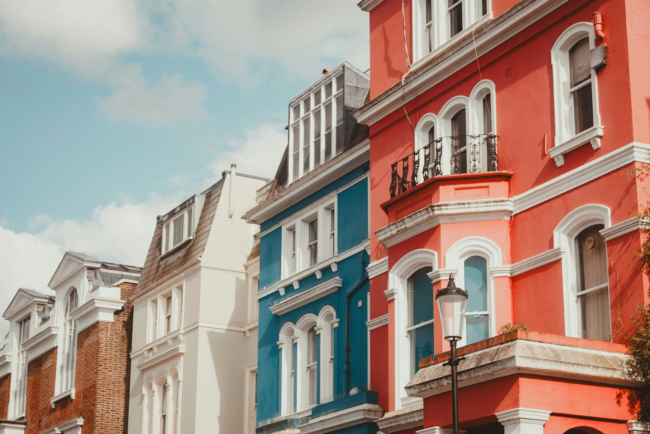 Colorful row of Victorian-style houses painted in white, blue, and red with ornate window frames and decorative trim, under a partly cloudy sky.