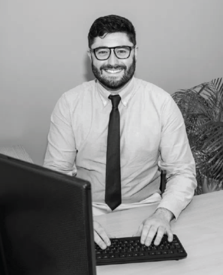 A man with glasses and a beard sitting at a desk in front of a computer, smiling, with a potted plant in the background.