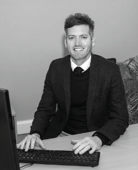 A man with curly hair and a beard sitting at a desk, smiling, with a computer keyboard and monitor in front of him, and a plant in the background.