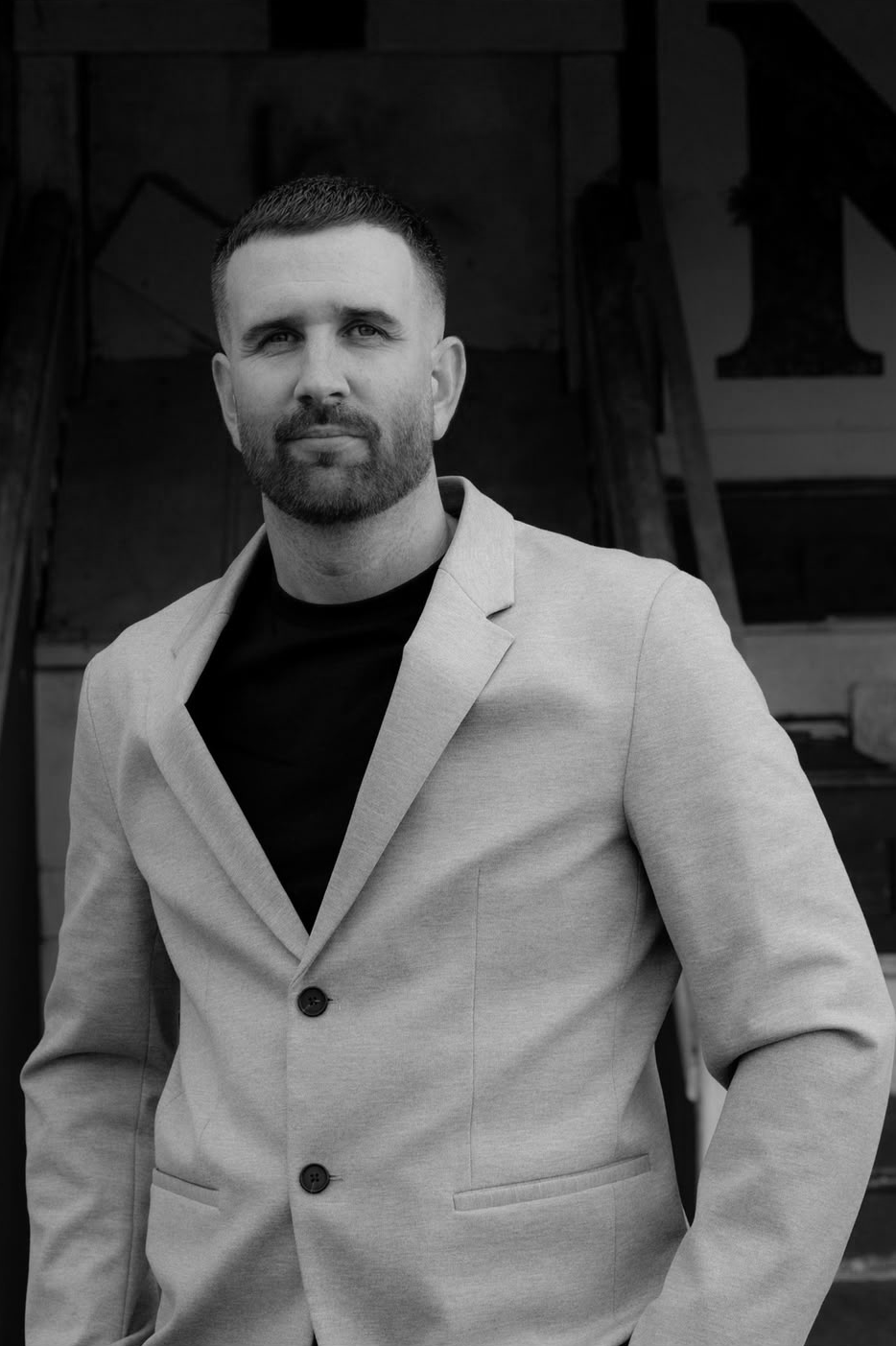 A man with short hair and a beard wearing a light blazer over a dark shirt, standing outdoors in front of a rustic wooden building.