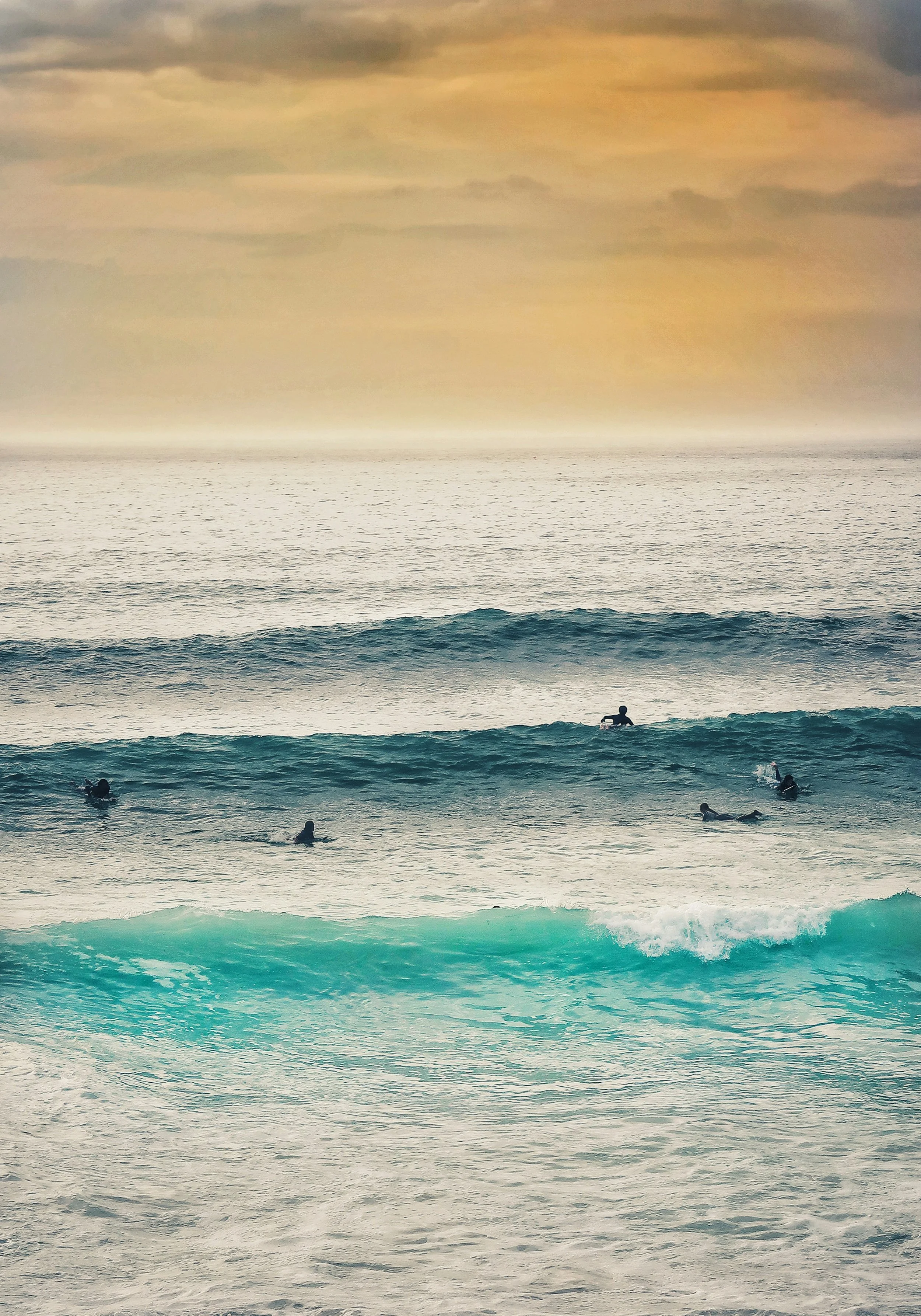 Several surfers waiting in the ocean near a sandy beach during sunset, with a cloudy sky overhead.