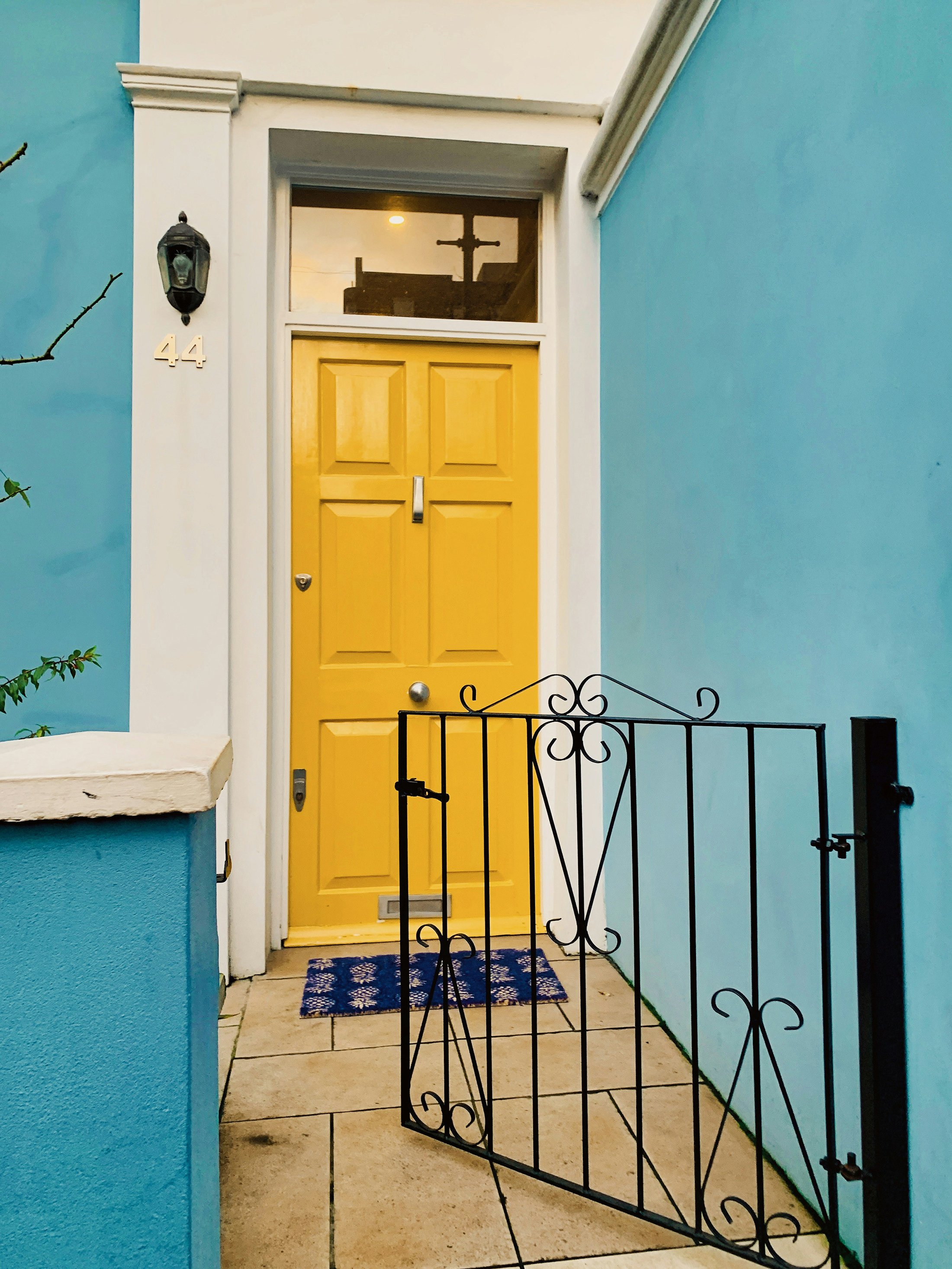 View of a house front door painted in yellow, with a black iron gate in front, blue walls on either side, a white door frame, a small window above the door with a reflection of a cross and a street light, a wall-mounted lantern light on the left side, house number 44 on the wall, and a blue and white doormat on the tiled porch.