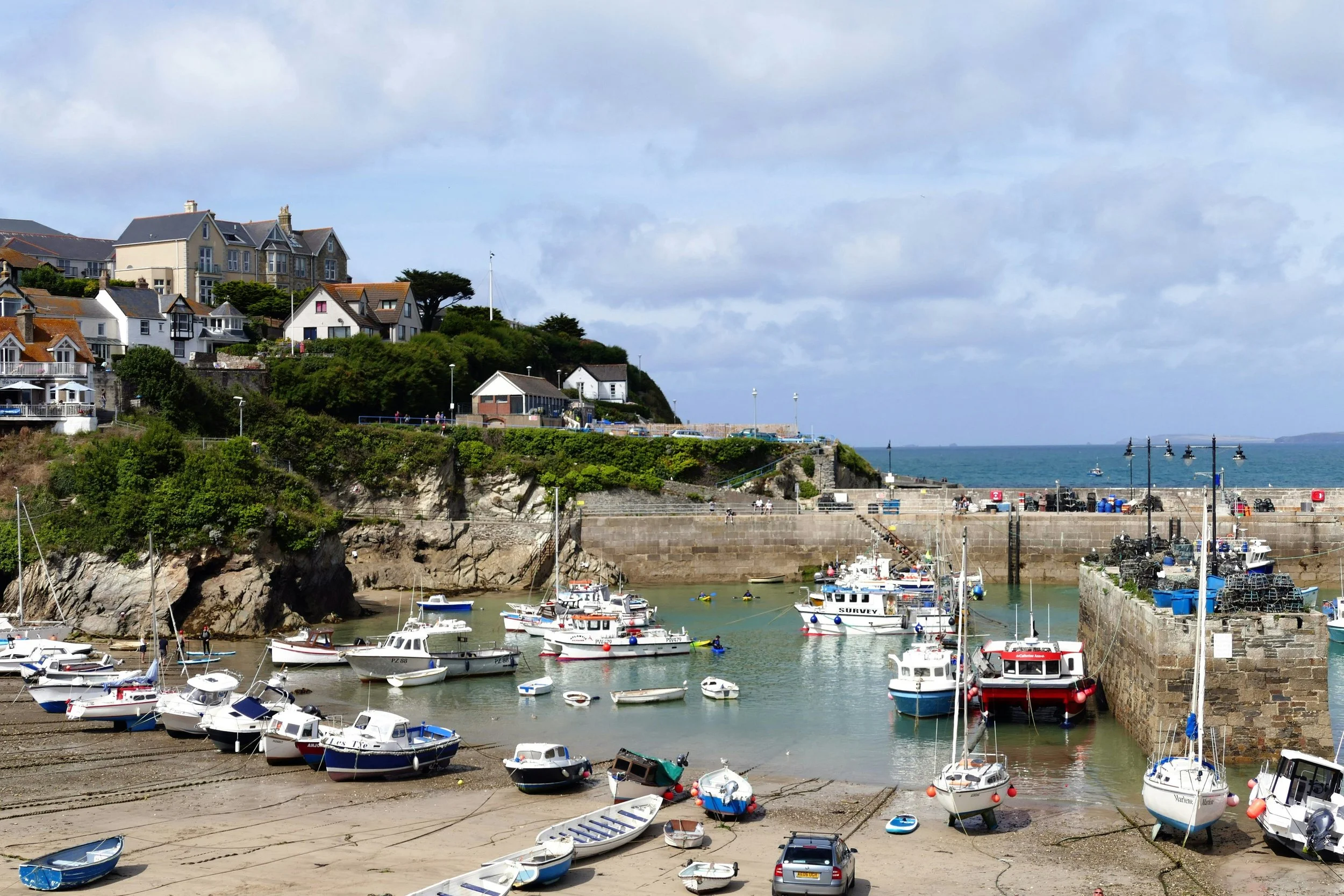 A coastal scene with a small harbor filled with boats, a stone pier, and a hillside with various houses and buildings overlooking the water. The sky is partly cloudy.
