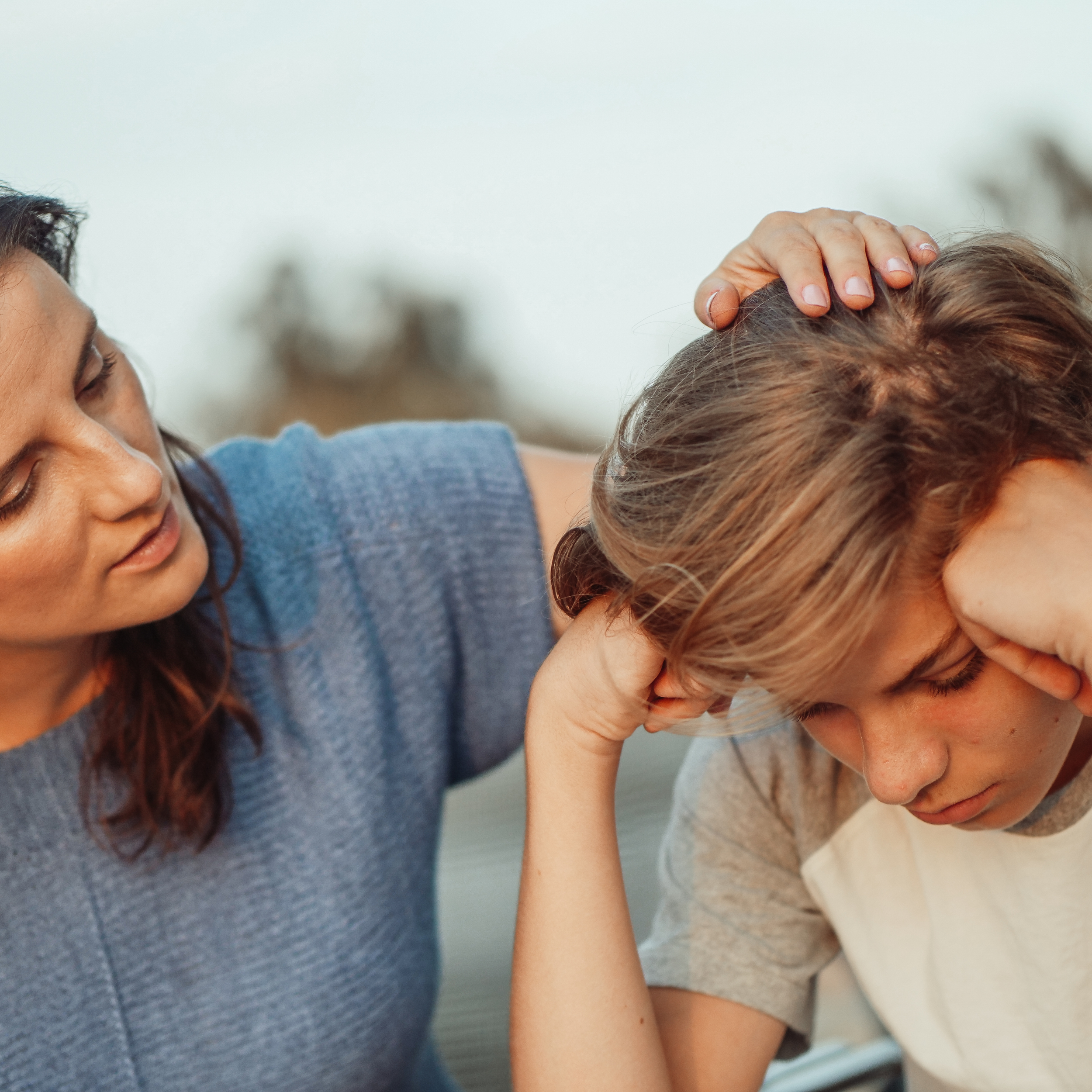 A person with reddish hair has their head bowed while another person, with dark hair and tan skin, places their hand gently on the other's head, possibly comforting or praying. The scene appears intimate and compassionate.