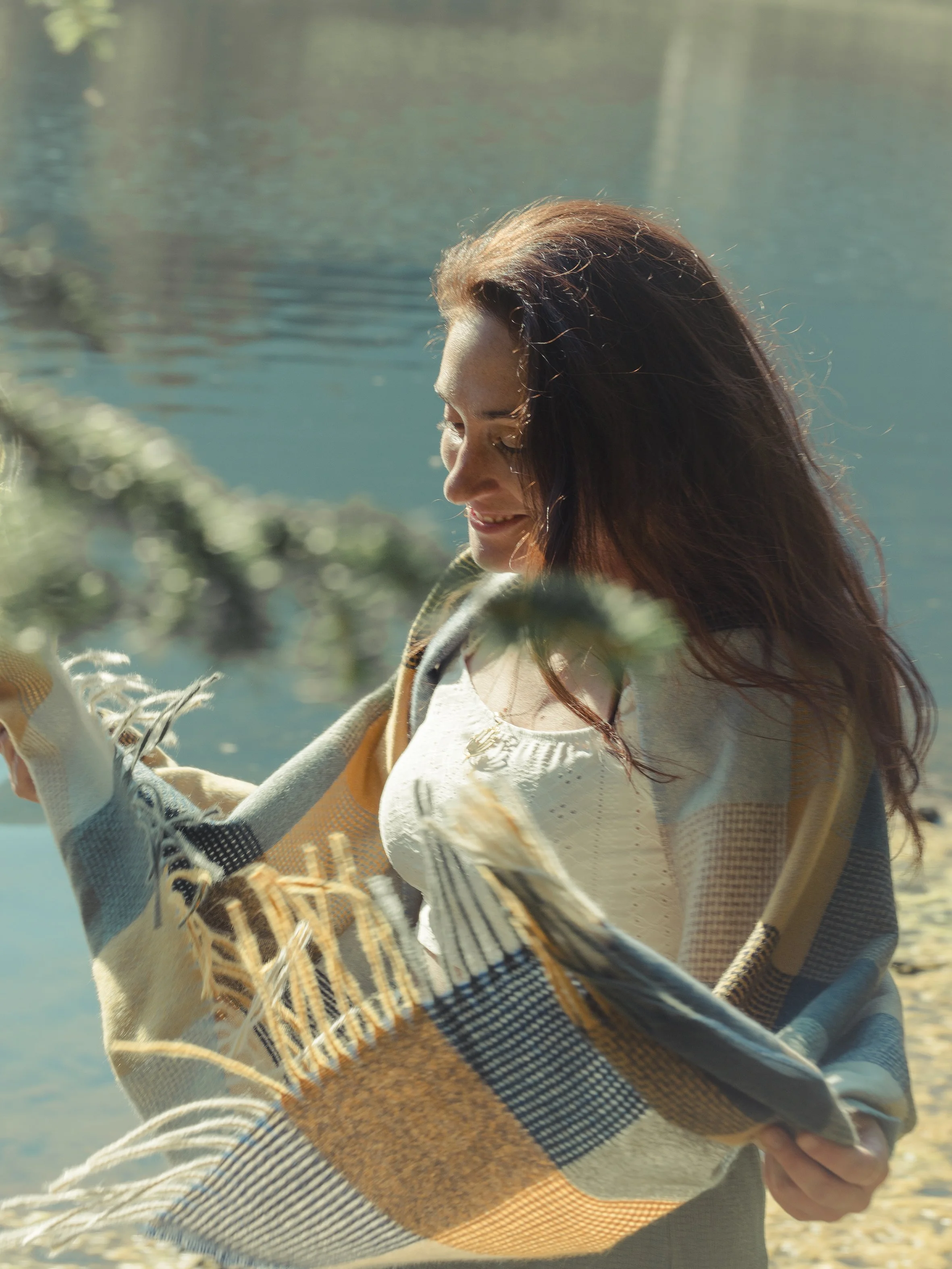 A woman with long brown hair sitting outdoors near a body of water, wrapped in a colorful, plaid blanket, smiling.