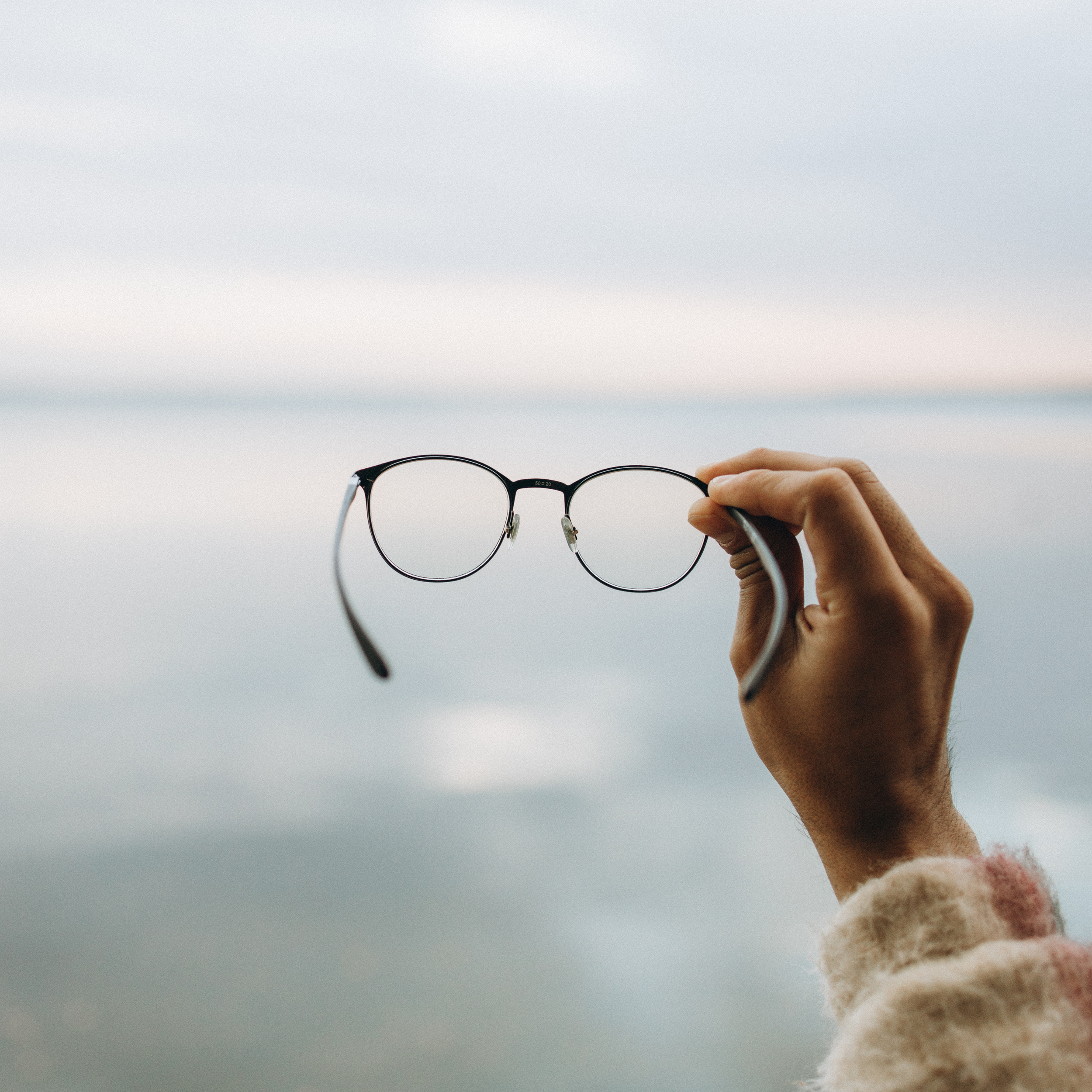 A person holding a pair of glasses up against a cloudy sky. The hand appears to be wearing a fuzzy, colorful sweater.