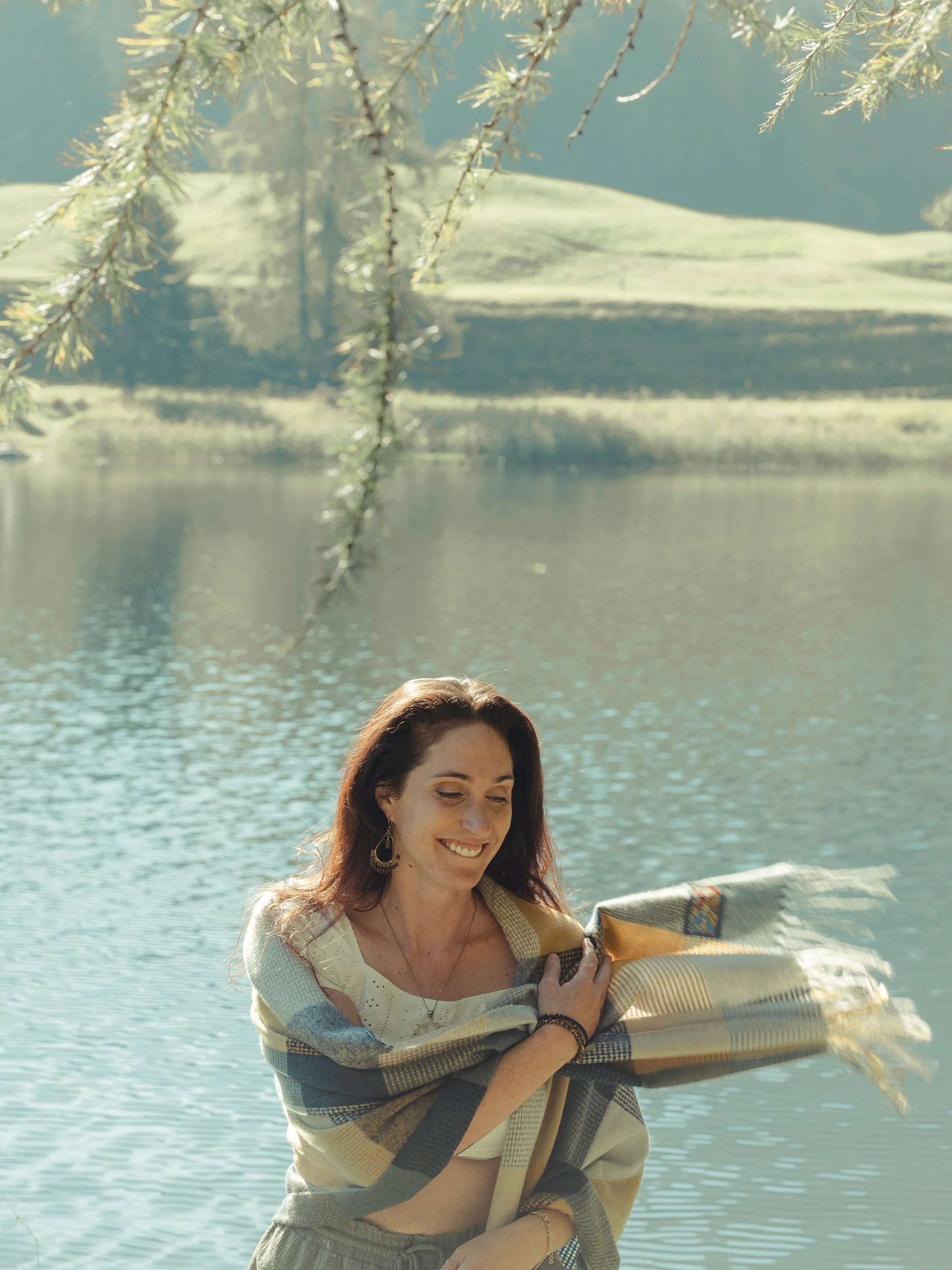 A woman with long brown hair and earrings smiles while holding a woven basket near a body of water with green hills in the background, under overhanging tree branches.