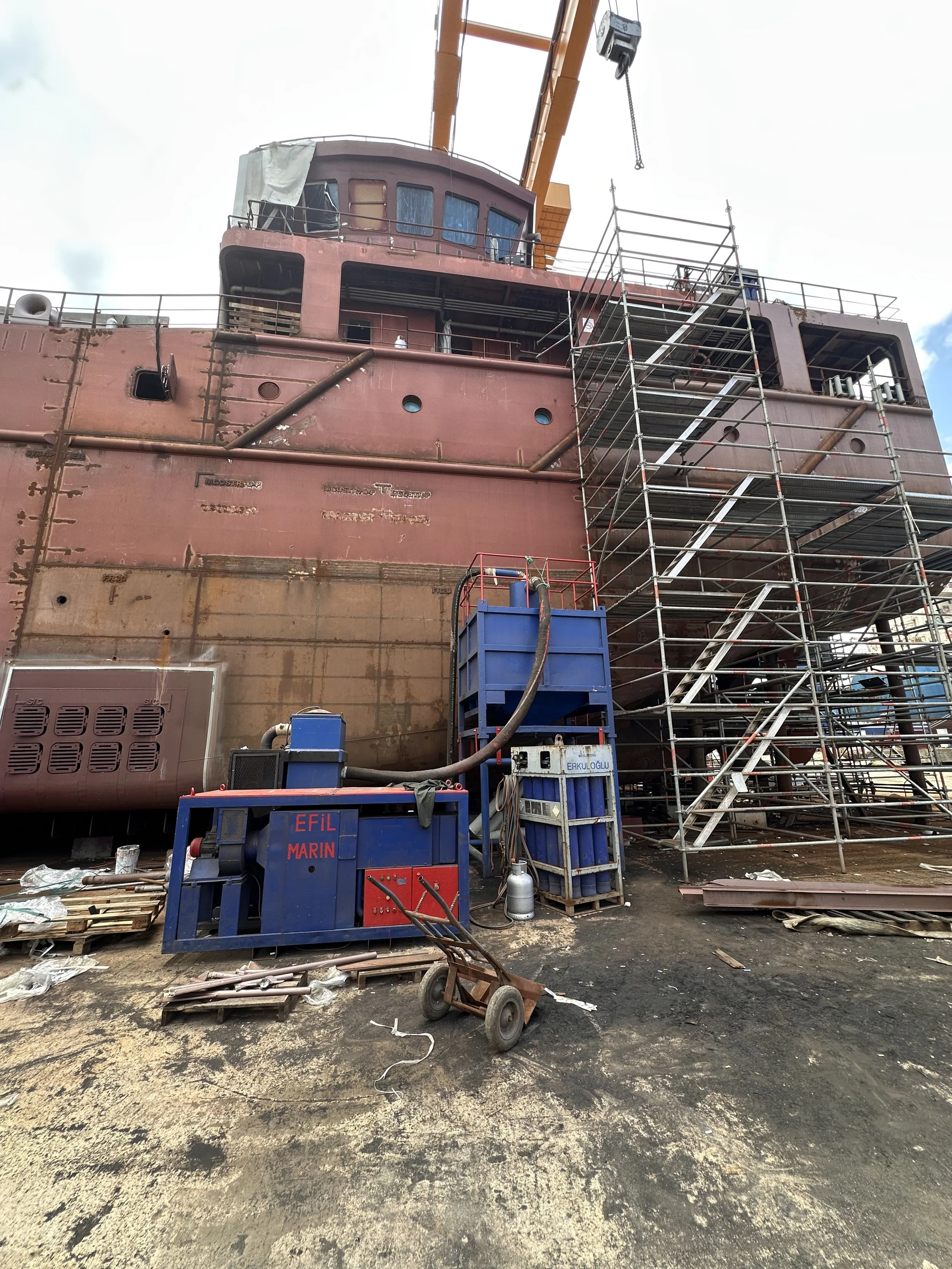 Yat limanı veya inşaat alanında gemi veya büyük makina yapım çalışmaları devam ediyor. Alanda çeşitli ekipmanlar, scaffolding ve malzemeler var. Yüksek yapılar, iş ekipmanları ve araçlar bulunuyor.