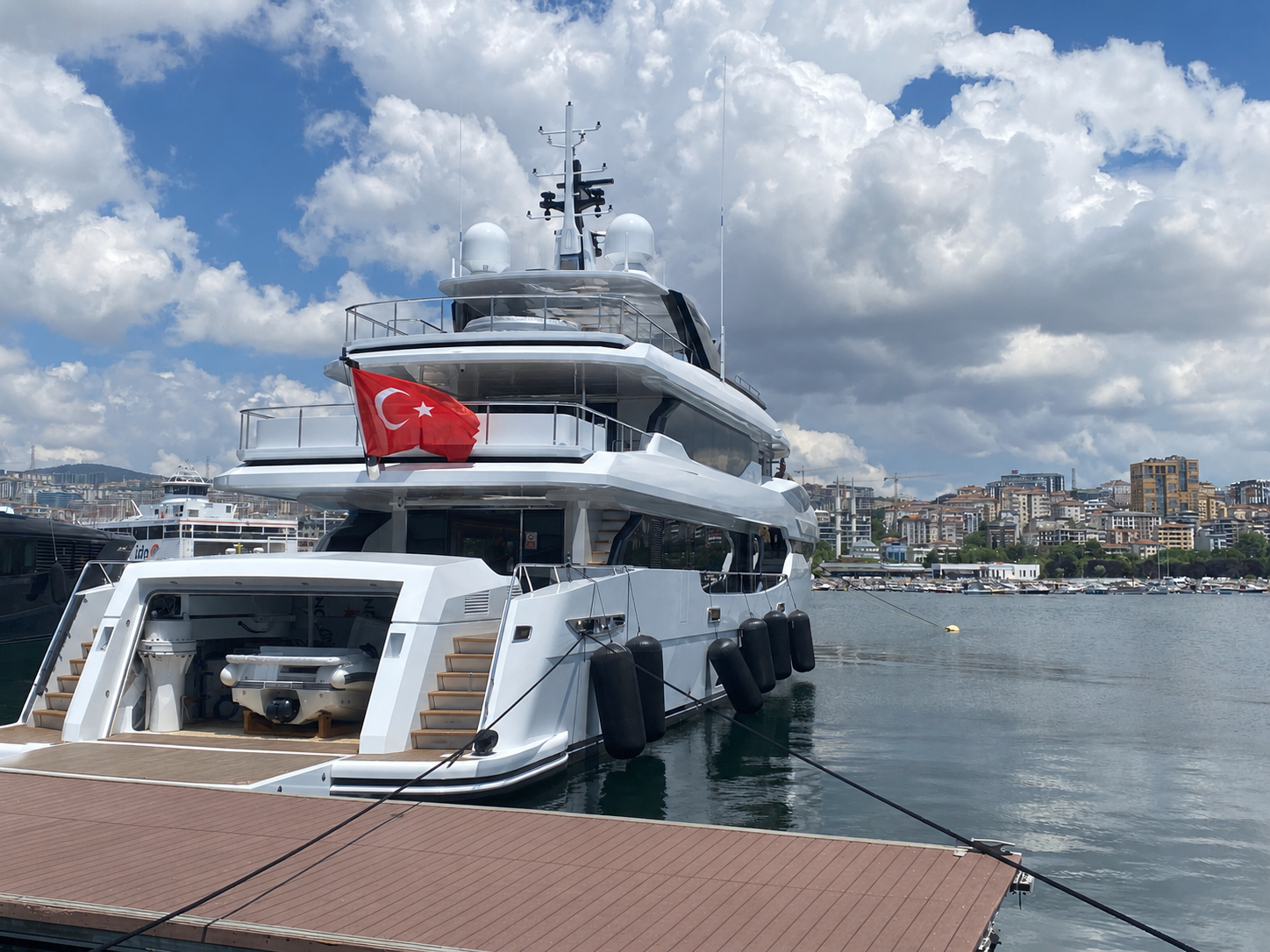 Bir yat denizde bağlı, Türk bayrağıyla süslenmiş, şehir manzarası ve bulutlu gökyüzü
