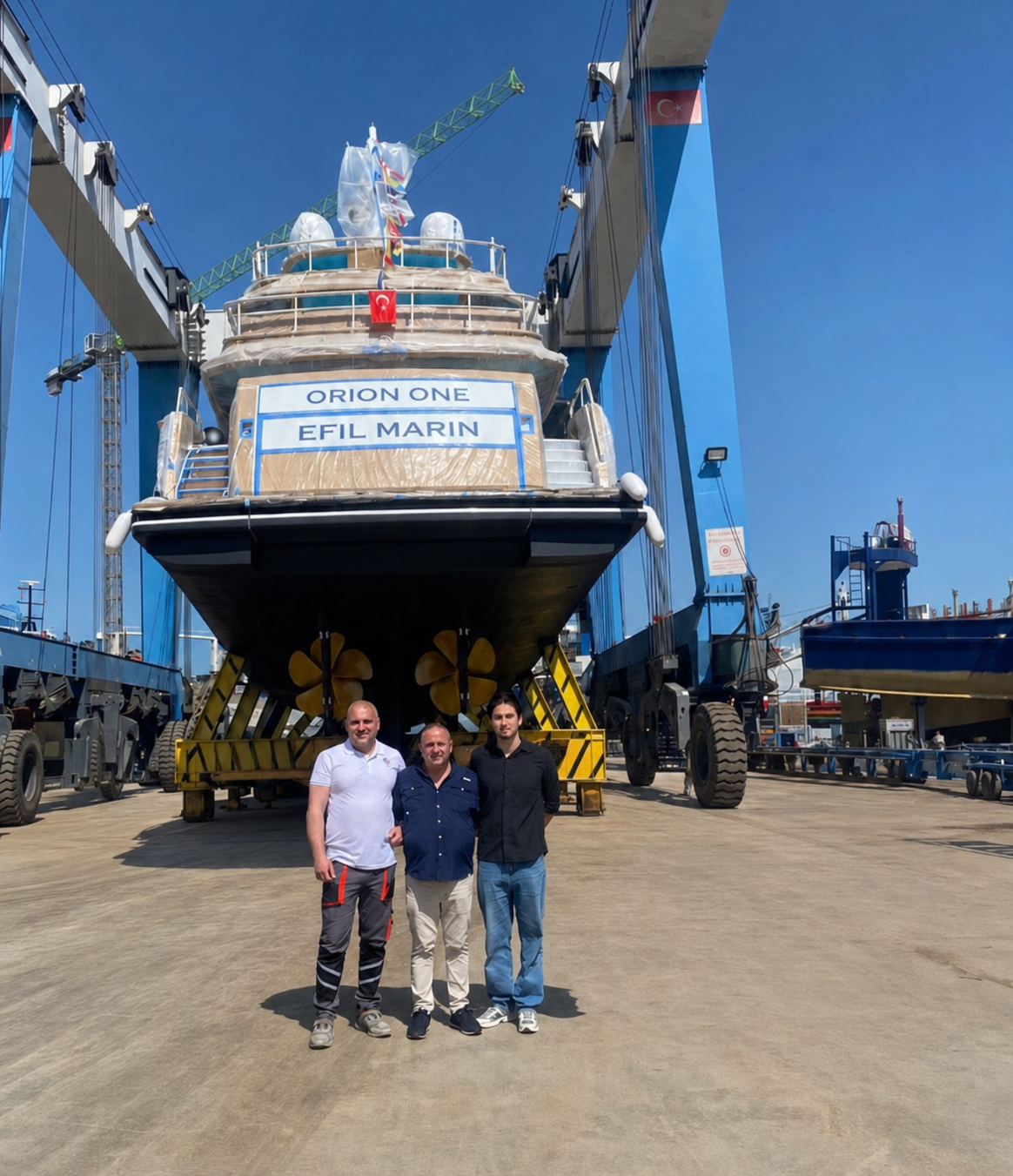 Üç adam, gökyüzü açık ve parlak, güneşli bir havada, deniz üzerinde inşa edilip bakıma alınmış büyük bir denizaltının önünde duruyor. Denizaltının üzerinde 'ORION ONE' ve 'EFIL MARIN' yazılı tabelalar var, ve arka planda vinç ve diğer tonajlı yapılar görülüyor.