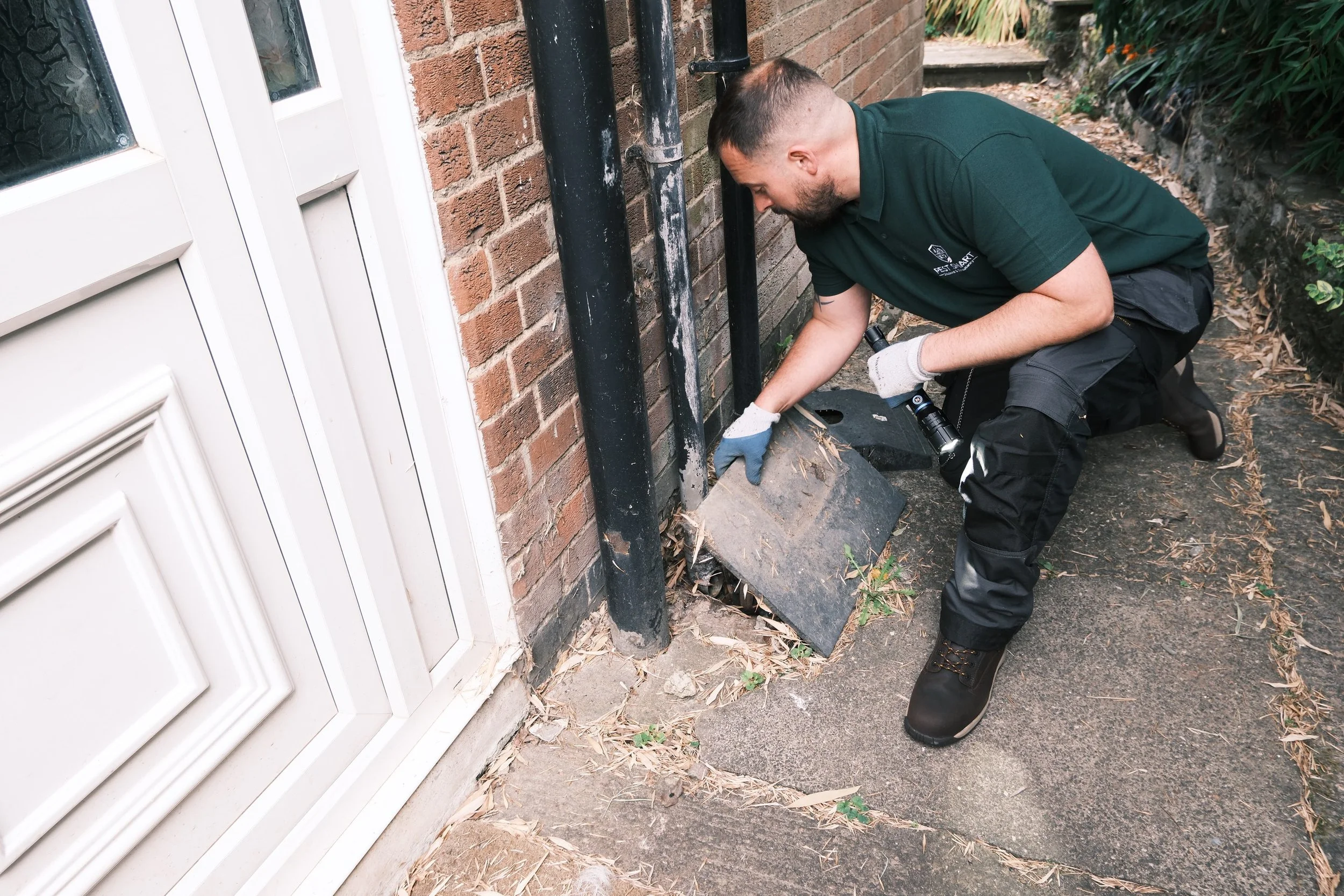 Man kneeling outside near a brick wall, working on a black pipe or conduit, wearing a green shirt, black pants, and gloves.