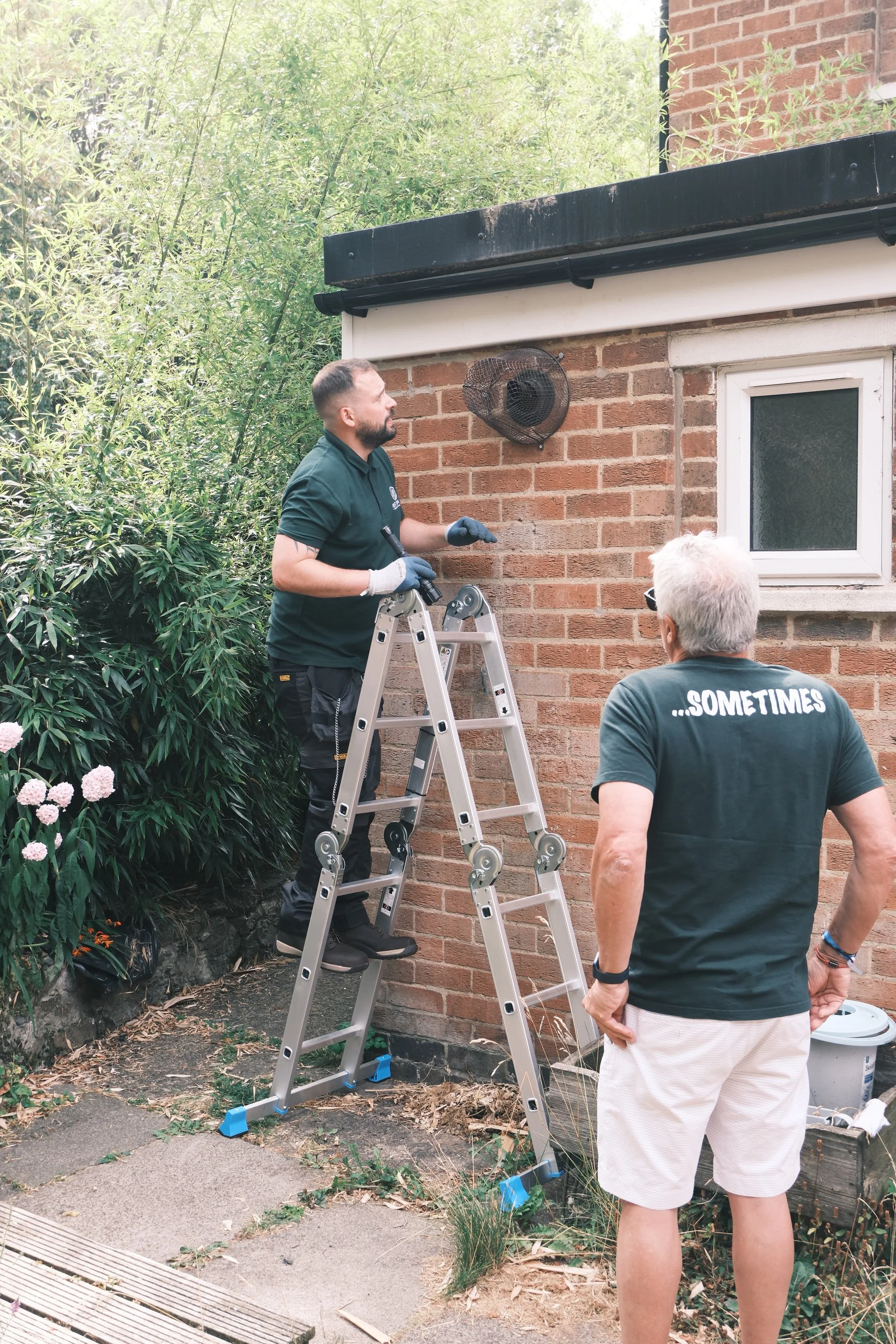 Two men working outside on a brick house. One man is on a ladder with gloves, pointing at something on the house wall. The other man is standing nearby with sunglasses, wearing a black T-shirt that says '...SOMETIMES' on the back. There are plants and flowers around.