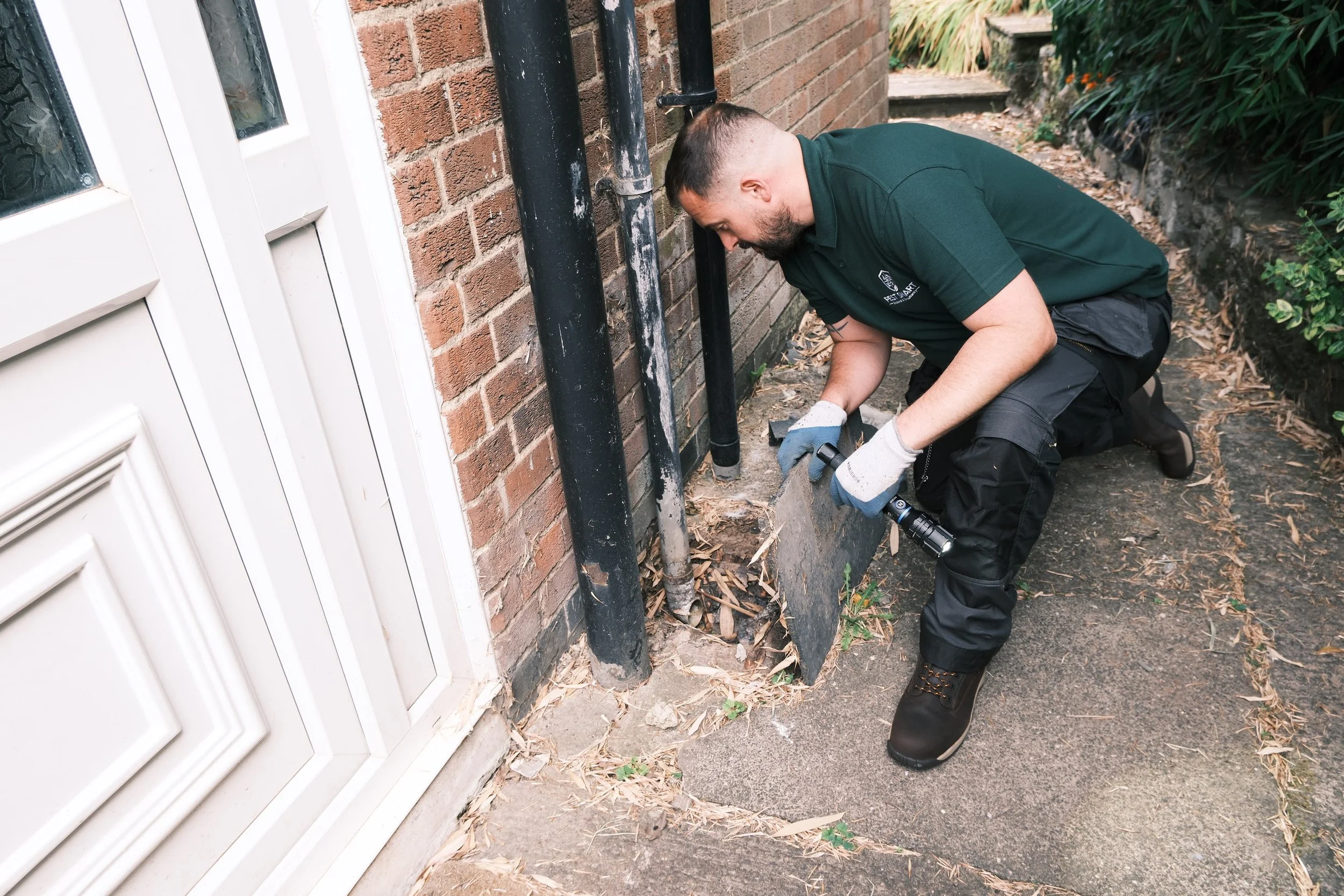 A man kneeling beside a brick wall, working on pipes and using a flashlight outside a house.