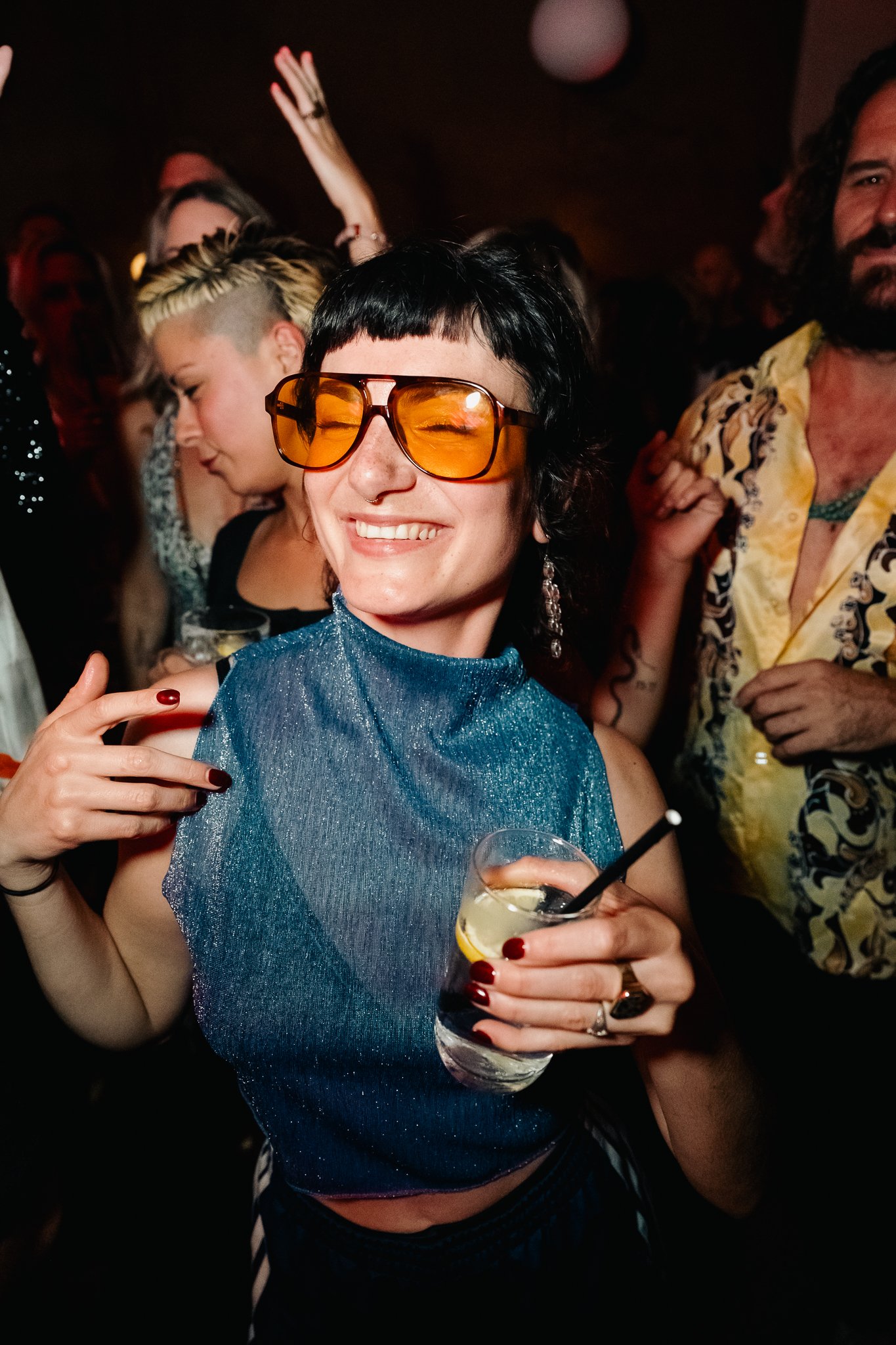 A woman with short black hair, wearing orange-tinted sunglasses and a shiny blue top, is smiling and holding a cocktail with lemon and cherry garnishes, at a lively party with other people dancing and enjoying themselves.
