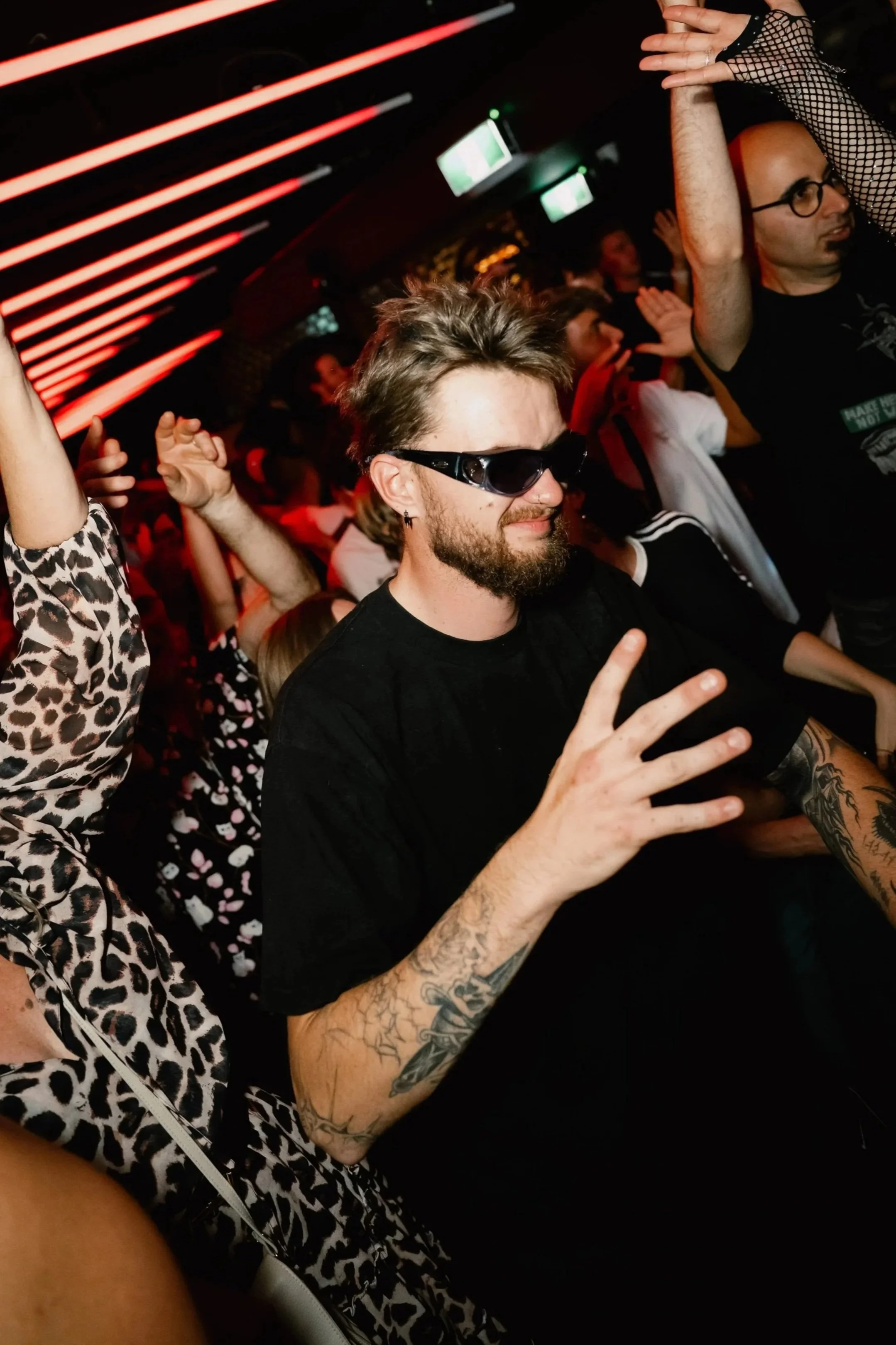People dancing at a party or club with red and green lighting, a man with a beard and sunglasses, and a woman with fishnet gloves.