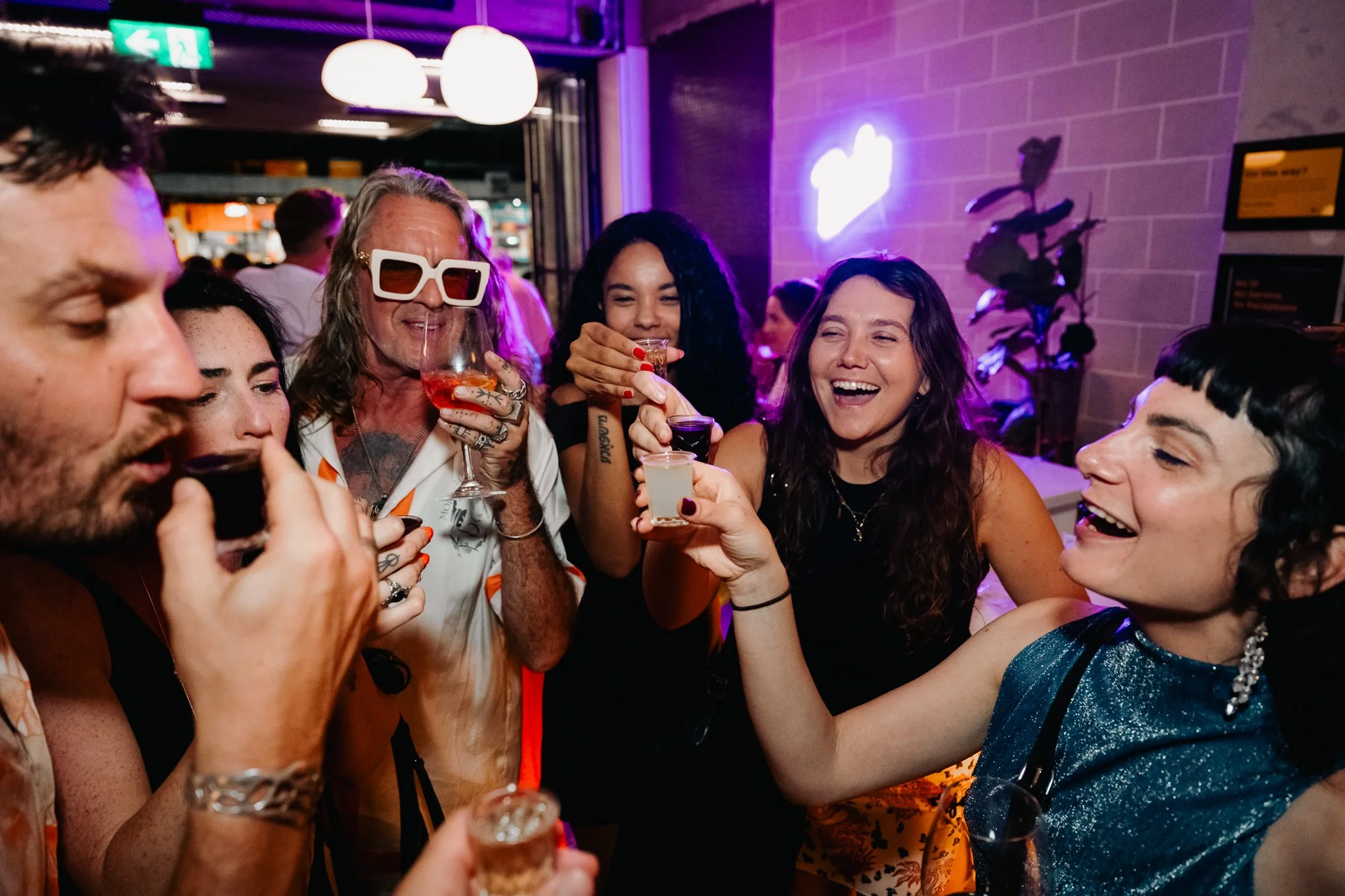 A group of people at a party are holding glasses and toasting, smiling, and laughing in a lively indoor venue with purple and pink lighting.