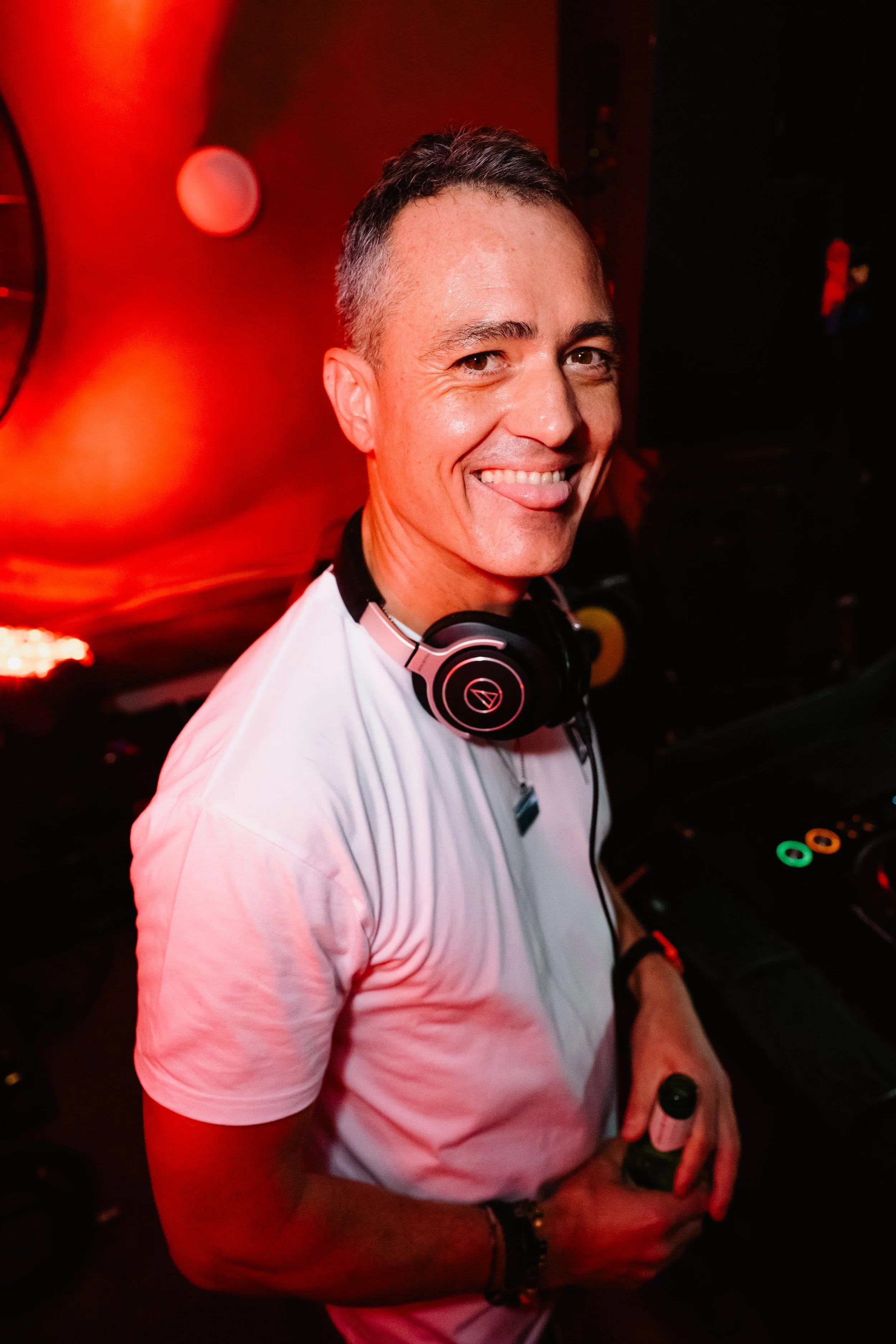 A man with short dark hair and a bright smile, wearing a white t-shirt, headphones around his neck, and holding a drink in a dark, red-lit environment, possibly a nightclub or party, with DJ equipment partly visible.