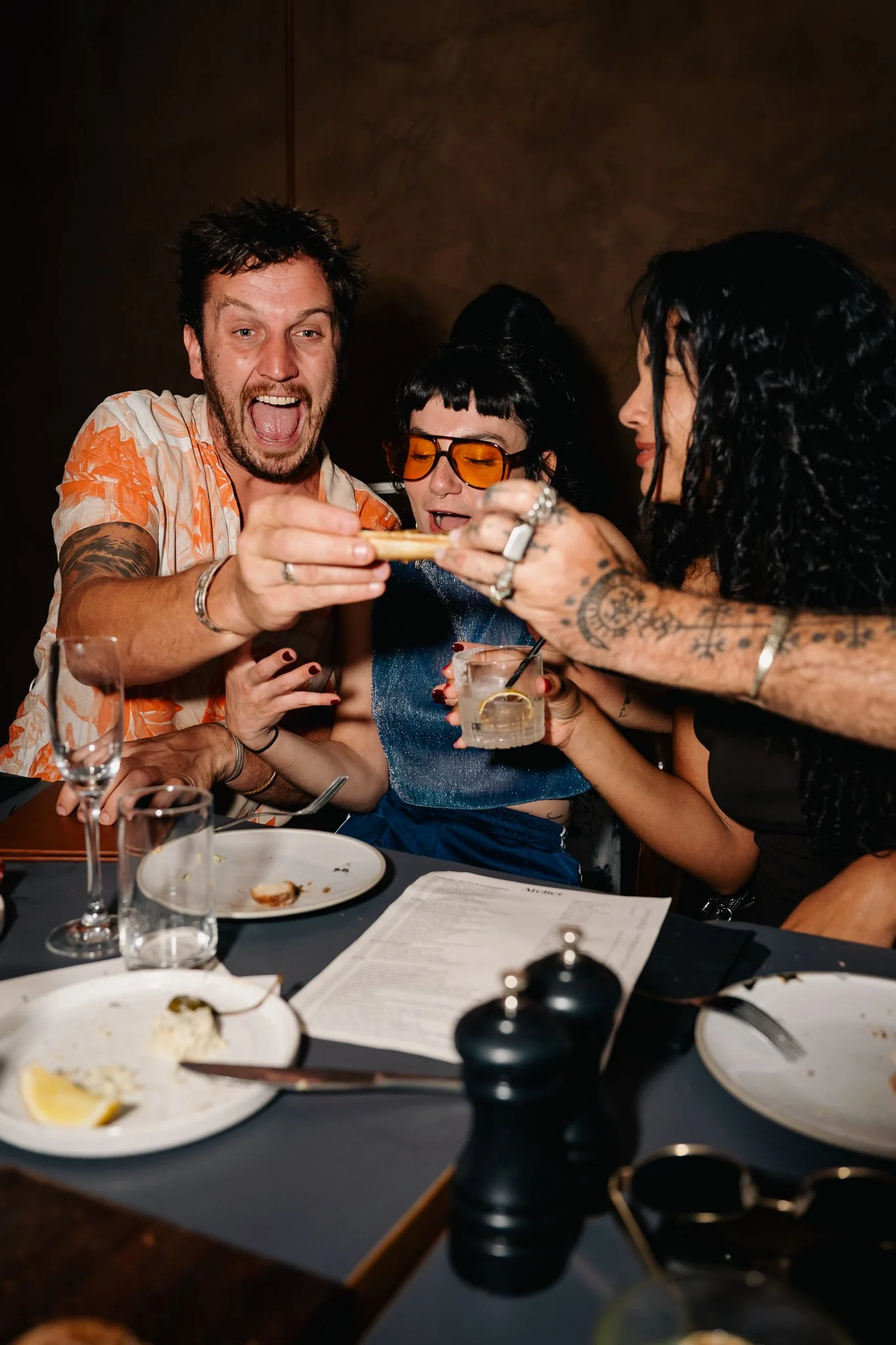 Three friends celebrating at a dinner table, with one holding a small dish, in a dimly lit restaurant.