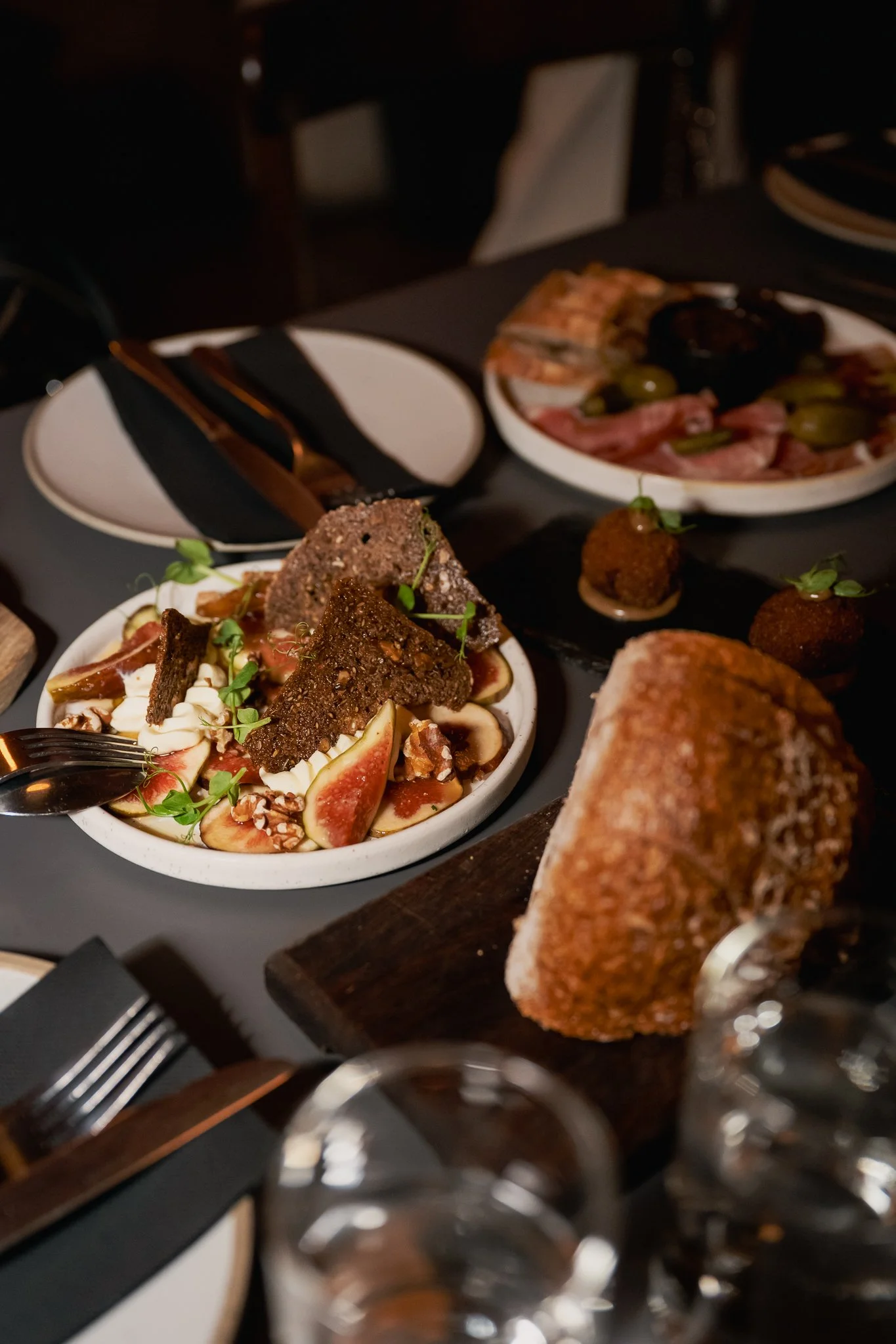 A dinner table with various dishes, including figs, bread, and appetizers, set with cutlery and glasses.
