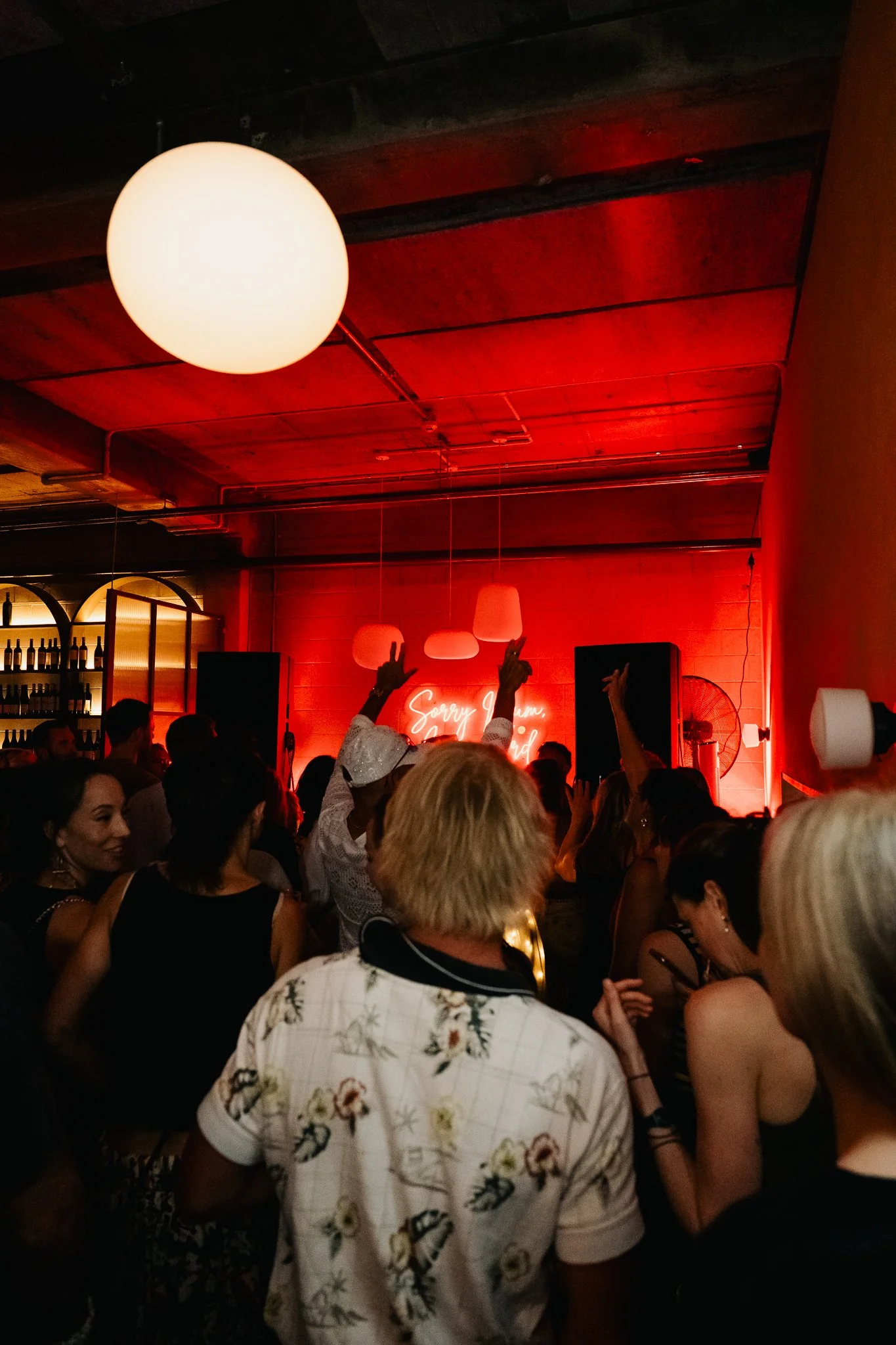 A lively indoor party scene with many people dancing and socializing. The room has red lighting, a neon sign that says 'Sorry, I'm...,' and modern hanging light fixtures. The ceiling is wooden, and there are some black and red elements visible.