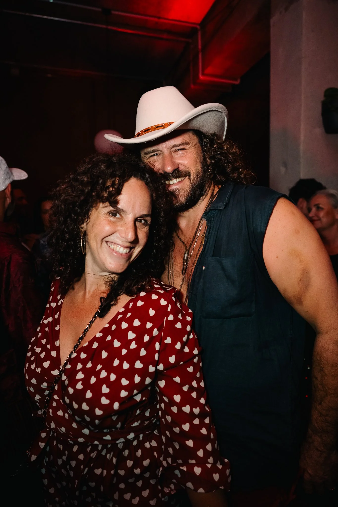 A man and woman smiling and posing closely together at a party or social event. The woman has curly dark hair and is wearing a red dress with white heart patterns. The man has long curly hair, a beard, and is wearing a white cowboy hat and a dark sleeveless shirt.