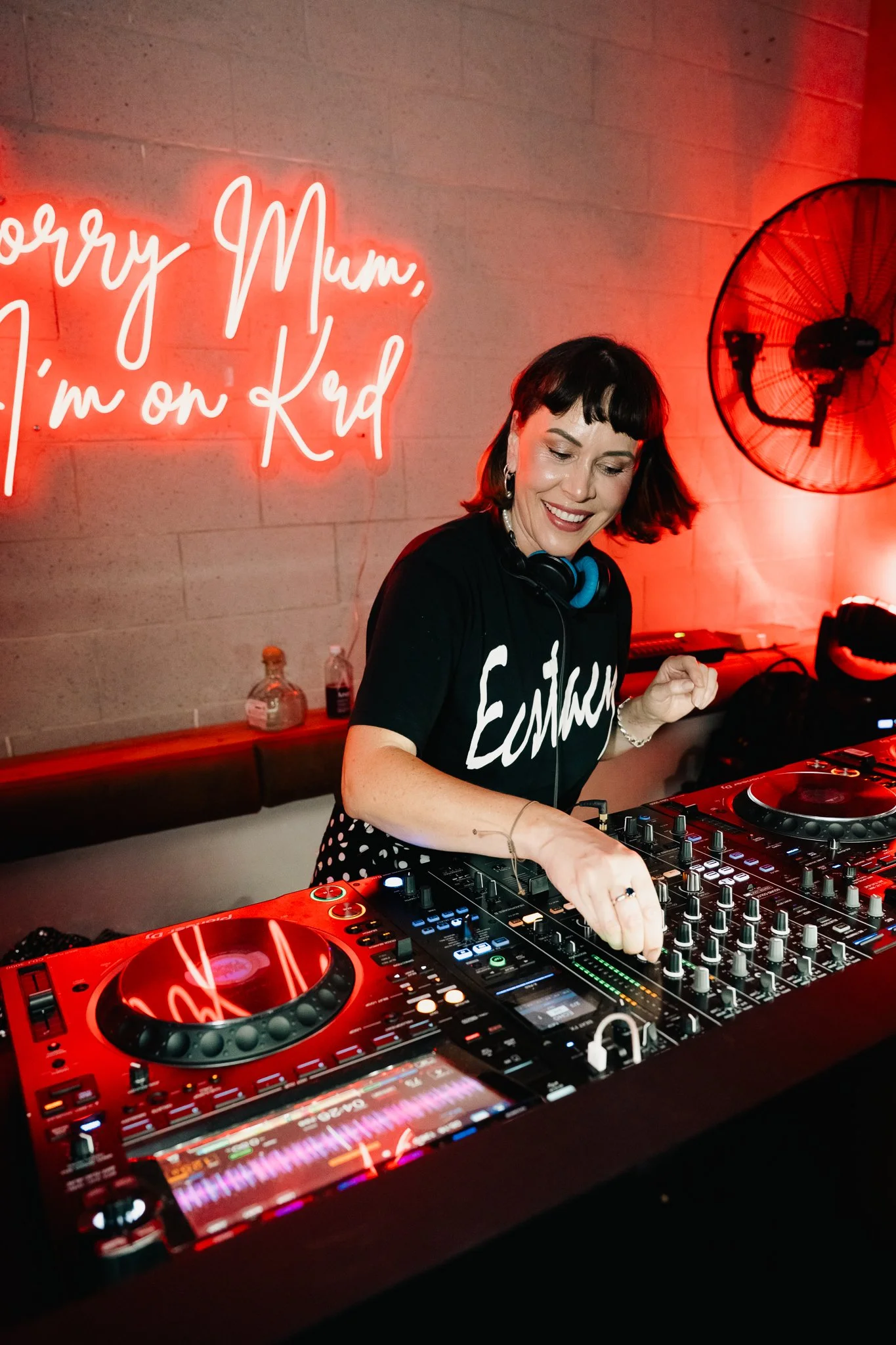 A woman DJ with short dark hair and bangs, smiling while wearing a black T-shirt with the word 'Euphoria' on it, is working on a DJ mixer with a red turntable in front. Behind her on the wall, there's a neon sign saying 'Sorry Mum, I'm on Krd' and a wall-mounted fan.