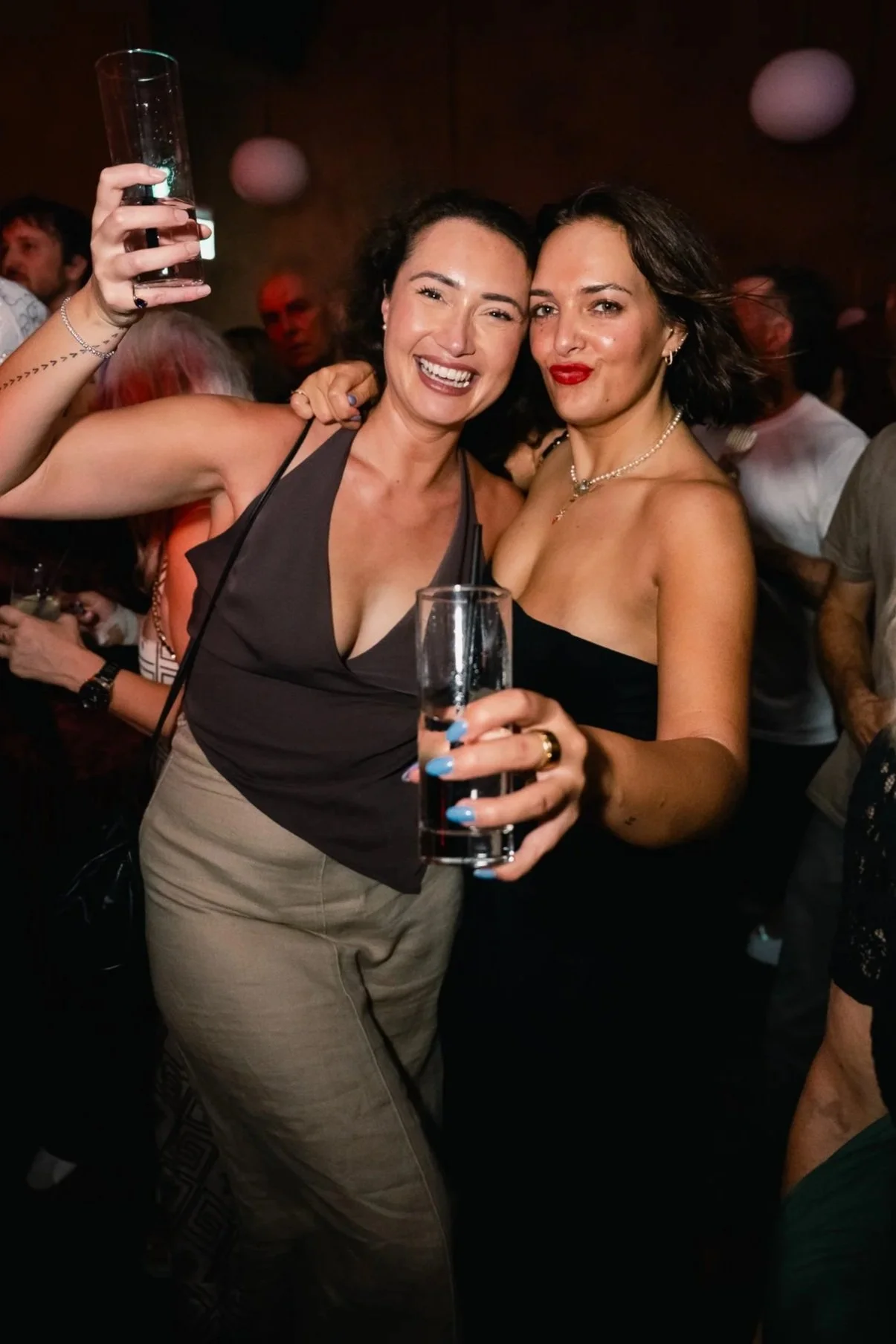 Two women celebrating at a party, smiling and holding glasses of drinks, with a dark background and other party-goers behind them.