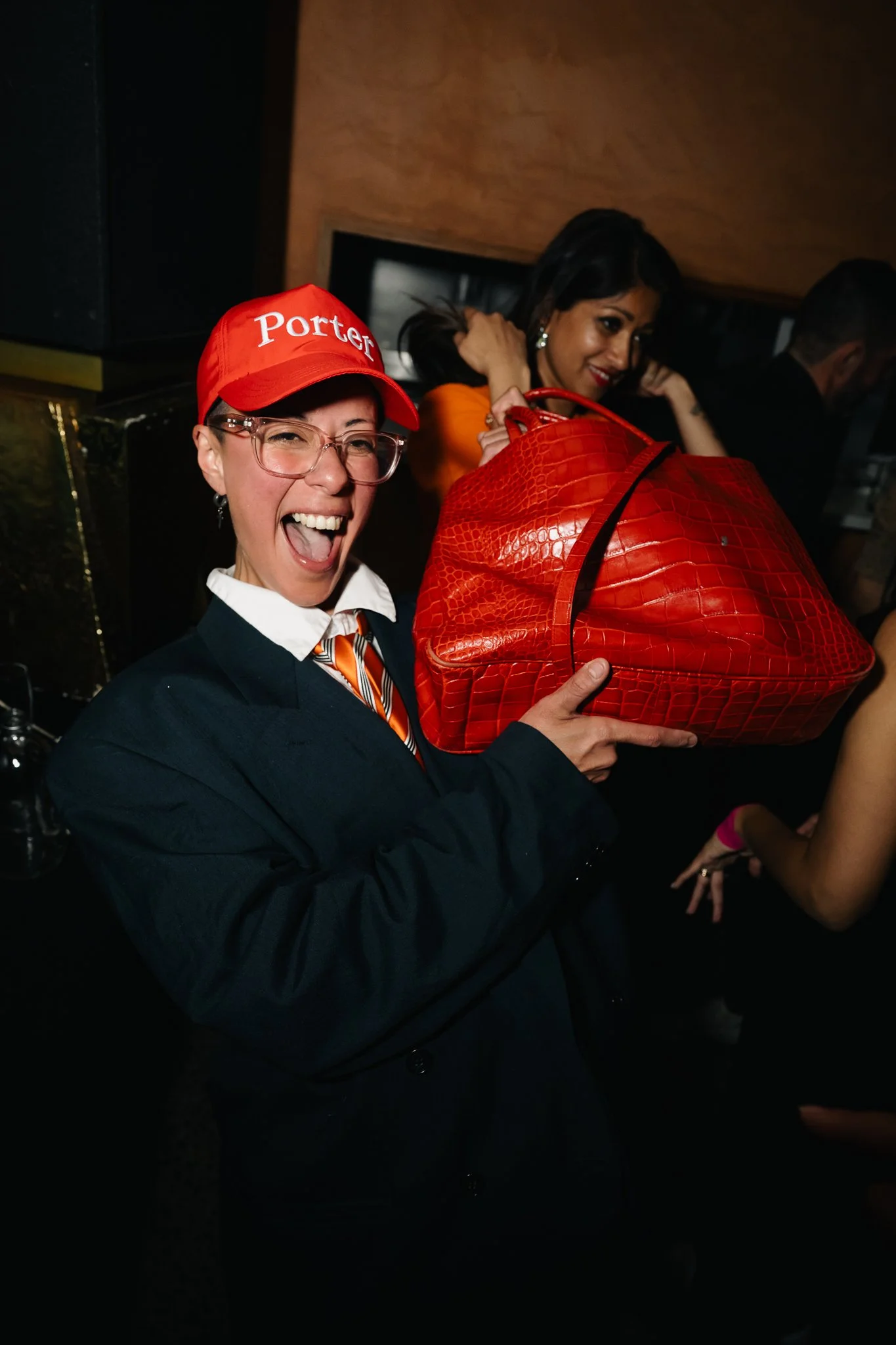 A woman in a dark suit and glasses is smiling widely and holding a large red handbag. She is wearing a red cap with 'Porter' written on it, and her outfit includes a white collar and a striped tie. Two other women are visible in the background, one a