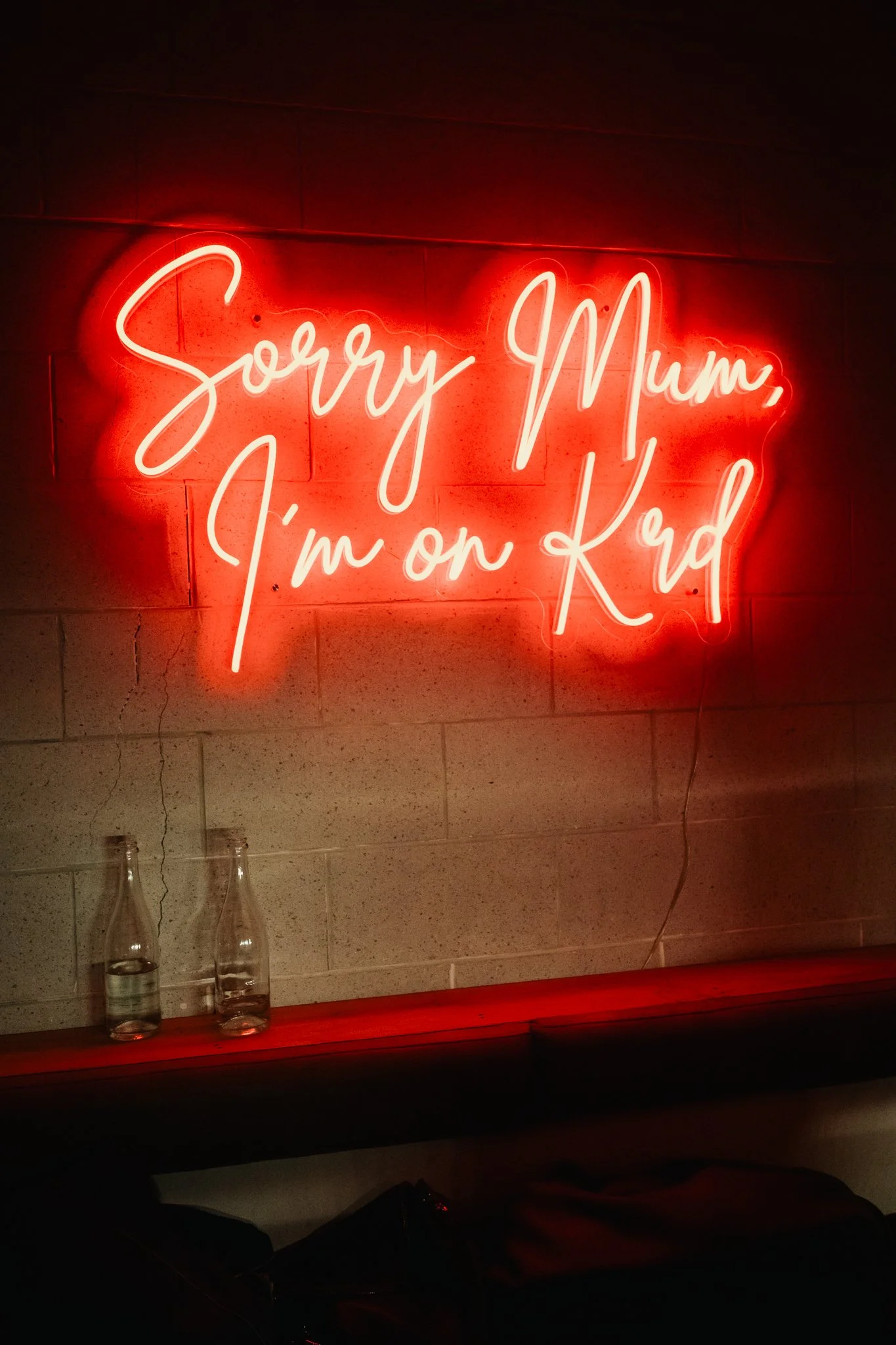 Neon sign on a wall that reads 'Sorry Mum, I'm on Kick' in cursive font, with two empty glass bottles below.