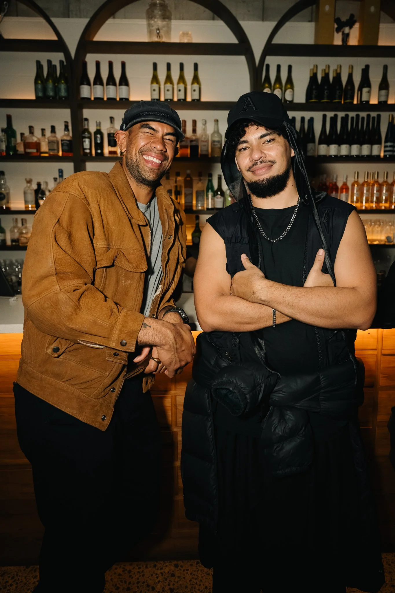Two men standing in front of a bar with shelves of liquor bottles, smiling and posing for the photo. One has a brown jacket and a black cap, the other has a black sleeveless shirt, a black cap, and a silver chain.