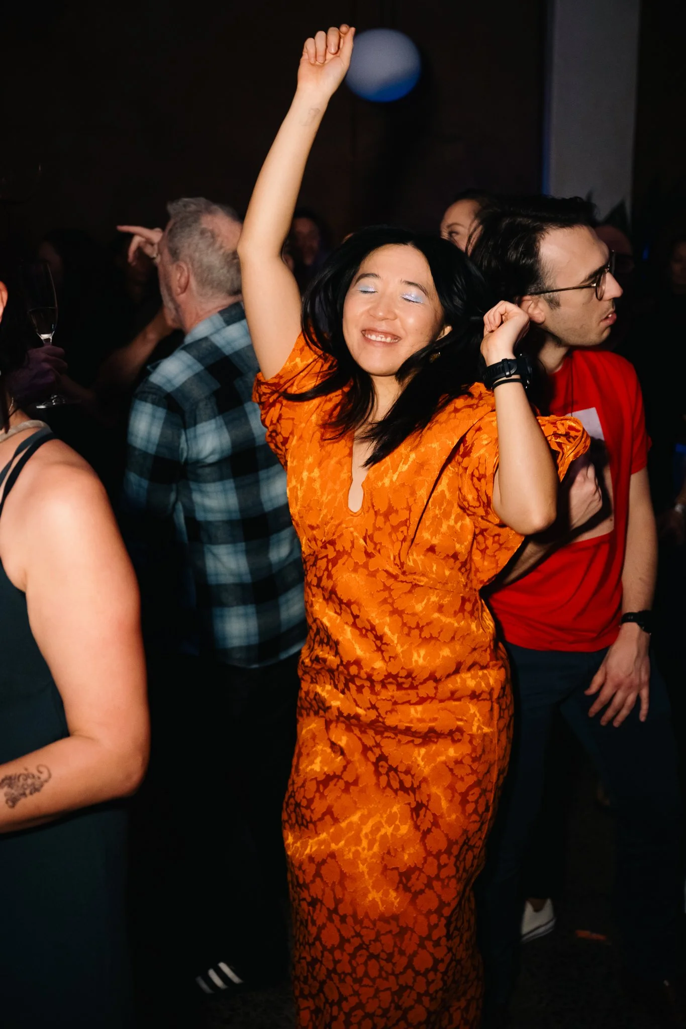 Woman in an orange patterned dress dancing with her eyes closed, smiling, and raising her arm at a party or nightclub while surrounded by other people.