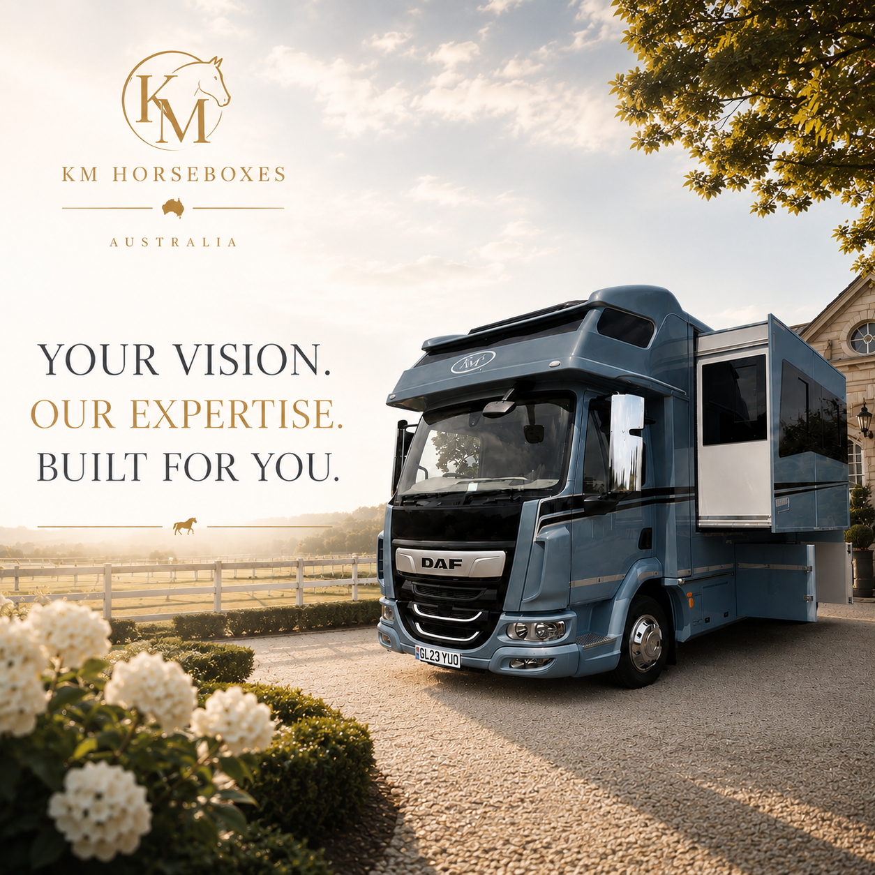 A large blue recreational vehicle parked on a driveway with manicured bushes and white flowers in the foreground, set against a background of a white picket fence, a green field, and a house with a porch. The sky is partly cloudy with sunlight shining through. The image includes a logo with a horse head and the text 'KM Horseboxes Australia,' and the slogan 'Your vision. Our expertise. Built for you.'