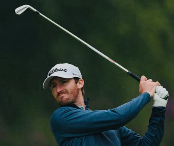Male golfer wearing a white Titleist cap and a blue jacket, holding a golf club after swinging on a golf course with green trees in the background.